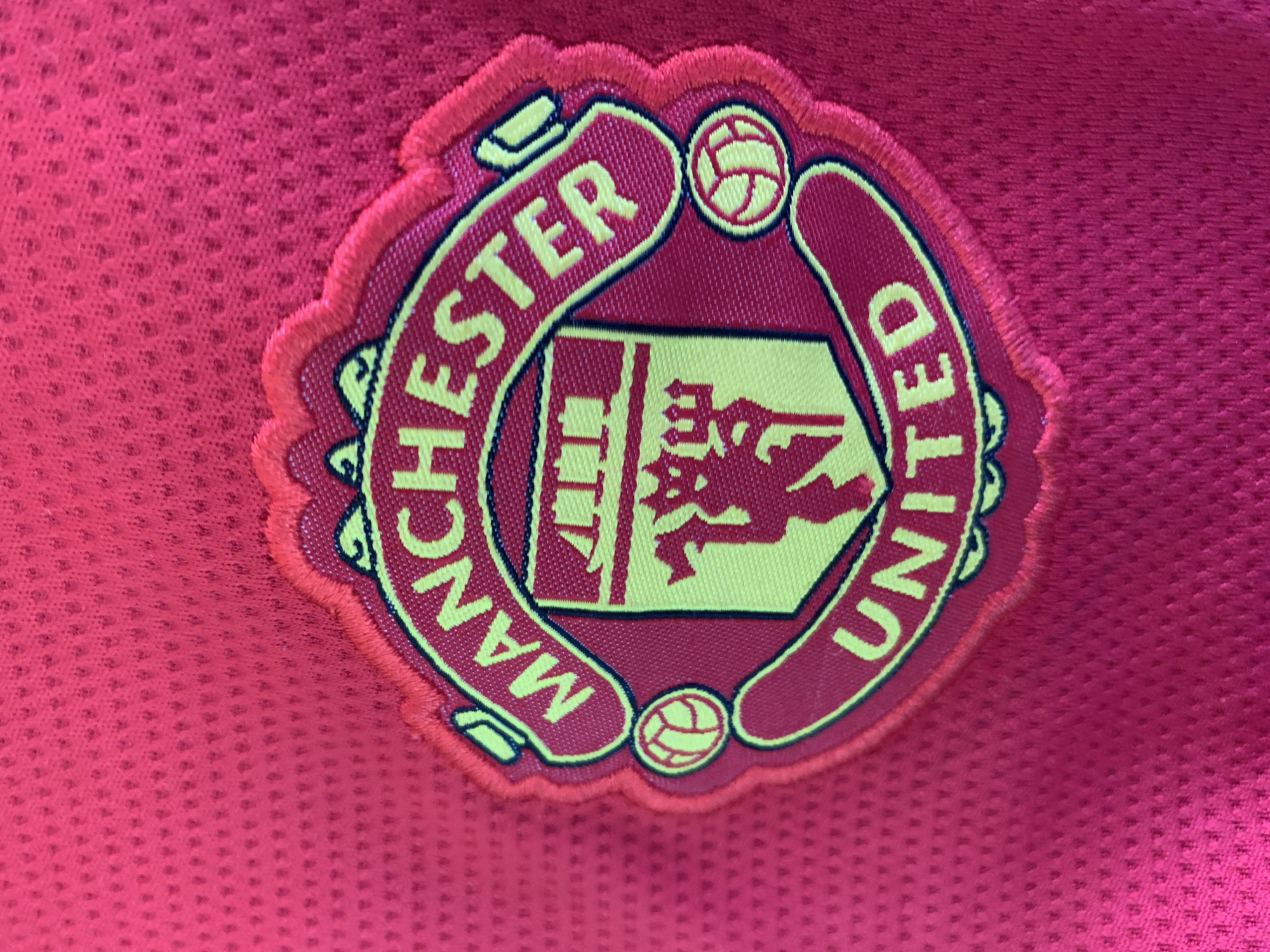 A close-up of a maroon Manchester United emblem is displayed on a matching maroon jersey. The emblem features the words "Manchester" and "United" in yellow lettering arranged in a semi-circular fashion around a crest containing a yellow devil figure, tower, and two soccer balls. The scene is located on a stadium.