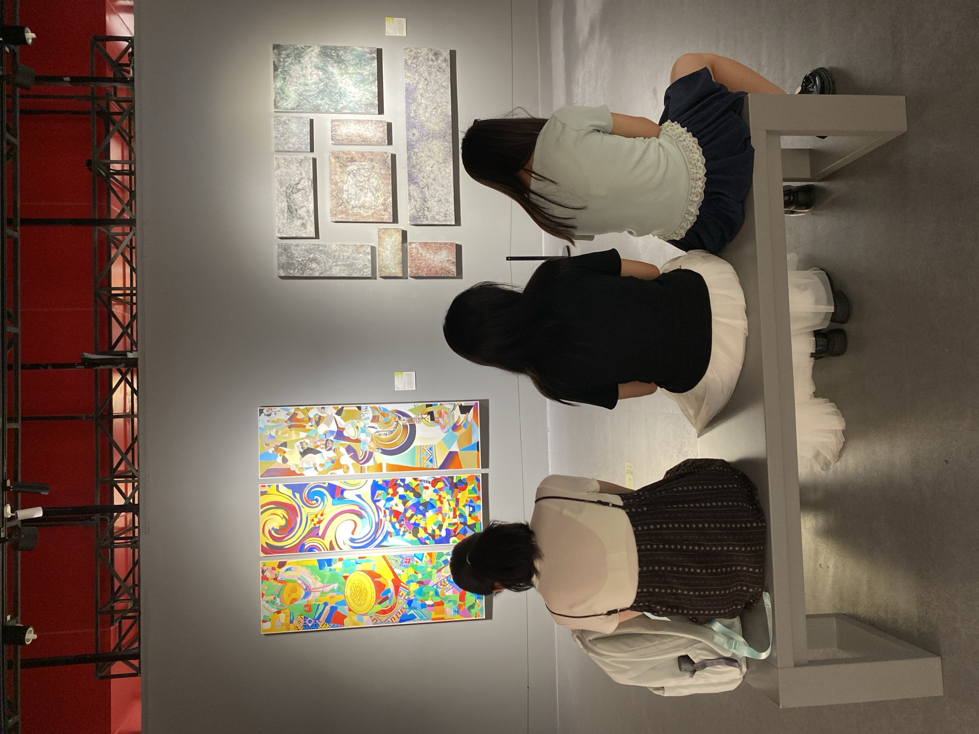 Three individuals are seated on a grey bench in what appears to be an art gallery, facing three colorful, abstract paintings on the left and a grid of textured, monochromatic artworks on the right. The lighting is dim, with spotlights focused on the artwork. The ceiling features industrial-style lighting fixtures and a bright red background structure. The floor is dark and polished. The individuals are viewed from behind, with their hair colors and clothing styles varying. The person on the left has dark hair tied back in a ponytail, is wearing a white top with a dark patterned dress, and has a light blue backpack next to them. The middle person has long, dark hair and is wearing a black shirt with a white skirt or dress. The person on the right has long, dark hair, wears a light blue t-shirt, and a dark blue skirt. They are all looking at the artwork. The presence of artwork displayed on a wall and individuals observing it strongly suggests an exhibition space. The question about the location being Yuzhong District, China, cannot be determined from the visual information provided in the image itself.