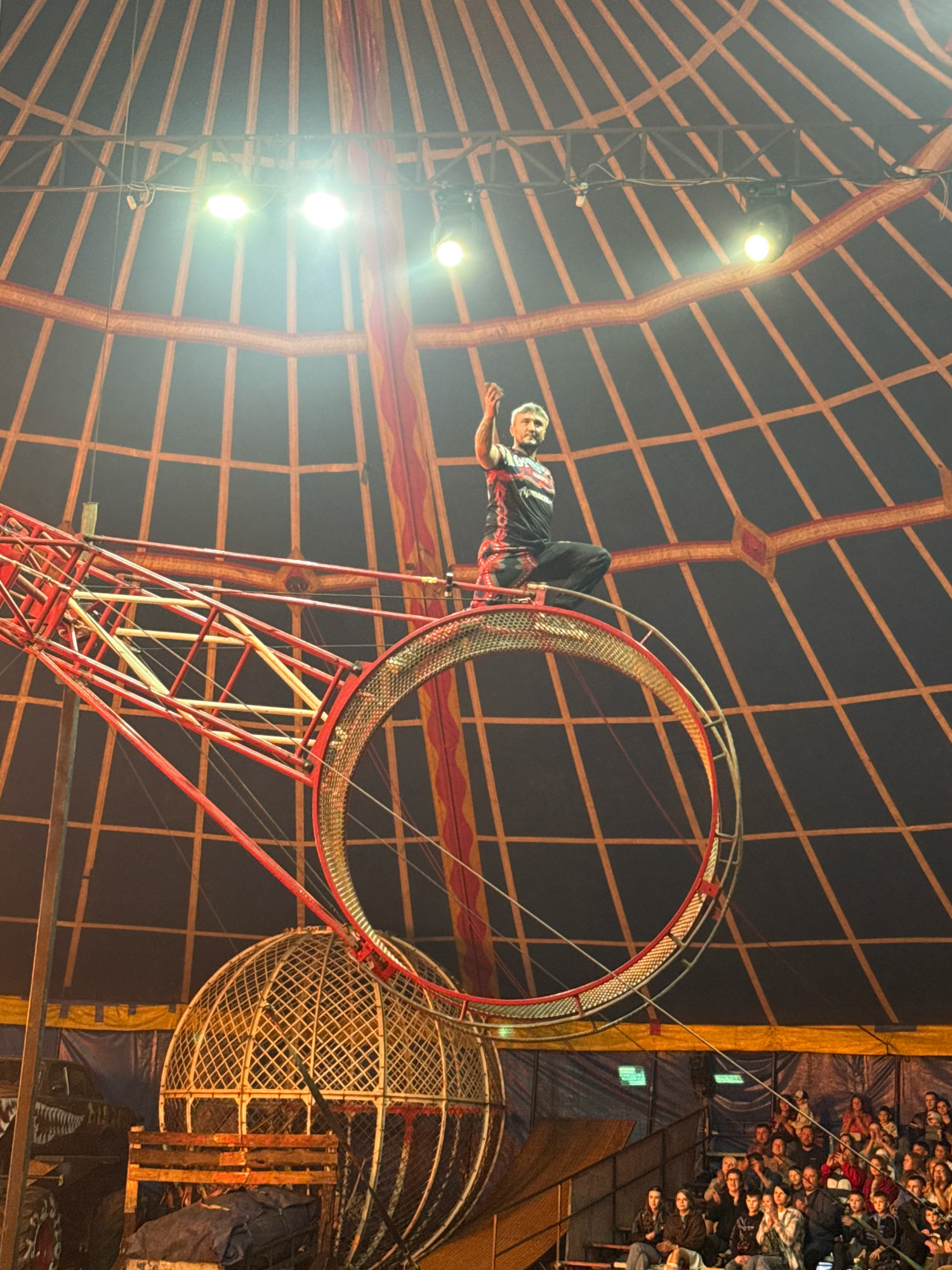 In Rusanovo, Russia, a performer is situated at the apex of a large metal rotating sphere within a circus tent. The performer, wearing a black and red patterned costume with a red collar, is seated with their right arm raised. The sphere, constructed with a red metal framework and a mesh-like interior, is partially visible. Below the sphere, a large crowd of spectators is seated in tiered seating, watching the performance. The circus tent features a dark ceiling with a grid-like pattern of orange lines, illuminated by bright spotlights.