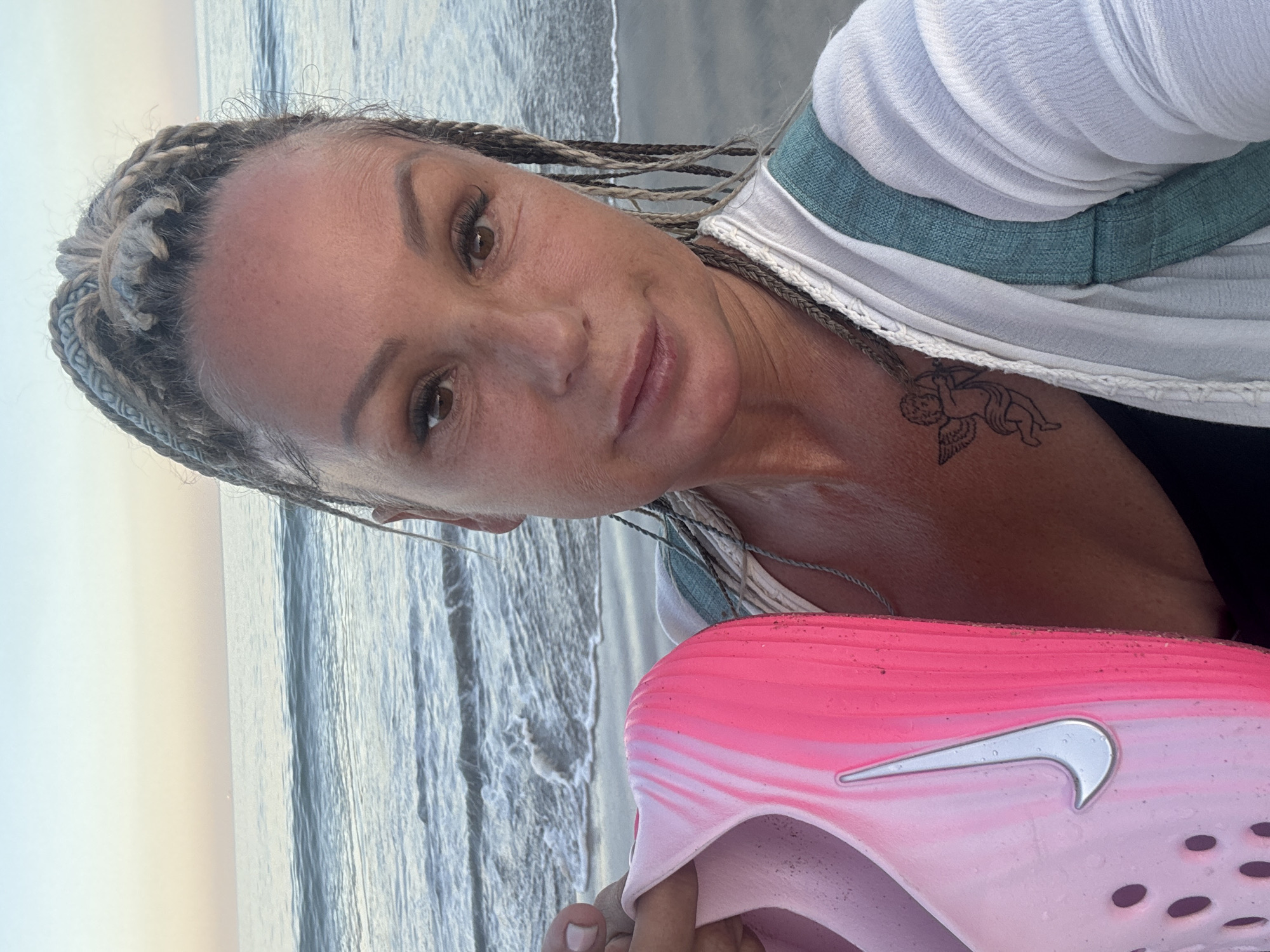 A woman takes a selfie on a beach in Kuta Selatan, Indonesia, with the ocean and sky forming the background. She has light-colored, possibly blonde or greyish, braids styled on her head and appears to be looking directly at the camera with a calm expression. Her eyes are brown, and she has visible eyelashes. She is wearing a white top with a teal/greenish band across the chest. A small tattoo of a cherub or cupid is visible on her right shoulder/neck area. In the lower right foreground, she holds a pink and white gradient sandal or slide shoe, which features a prominent silver Nike swoosh logo. The background shows the ocean with gentle waves, and the sky is a soft, light color, suggesting either dawn or dusk.