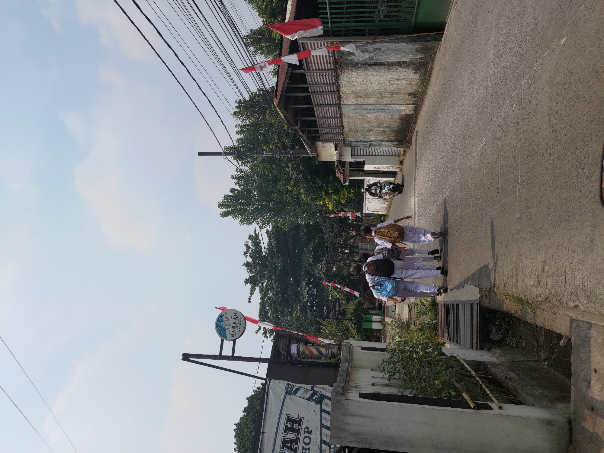 A clear, sunny day in Depok, Indonesia, is depicted as four students in white uniforms walk away from the camera down a paved road. The students, two with backpacks and two with bags, are walking in a group. A scooter rider is approaching them from the opposite direction. On the left side of the road, there is a barber shop with a sign featuring a barber pole and the word "BARKKAH." Further down the road, on the right, a building with a wooden porch and corrugated metal siding is visible. Several Indonesian flags, red and white, are displayed on poles along the street. The environment suggests a suburban or semi-urban area with residential and commercial elements. The road appears to be a local street with overhead utility lines. The drainage ditch on the left side of the road contains debris.