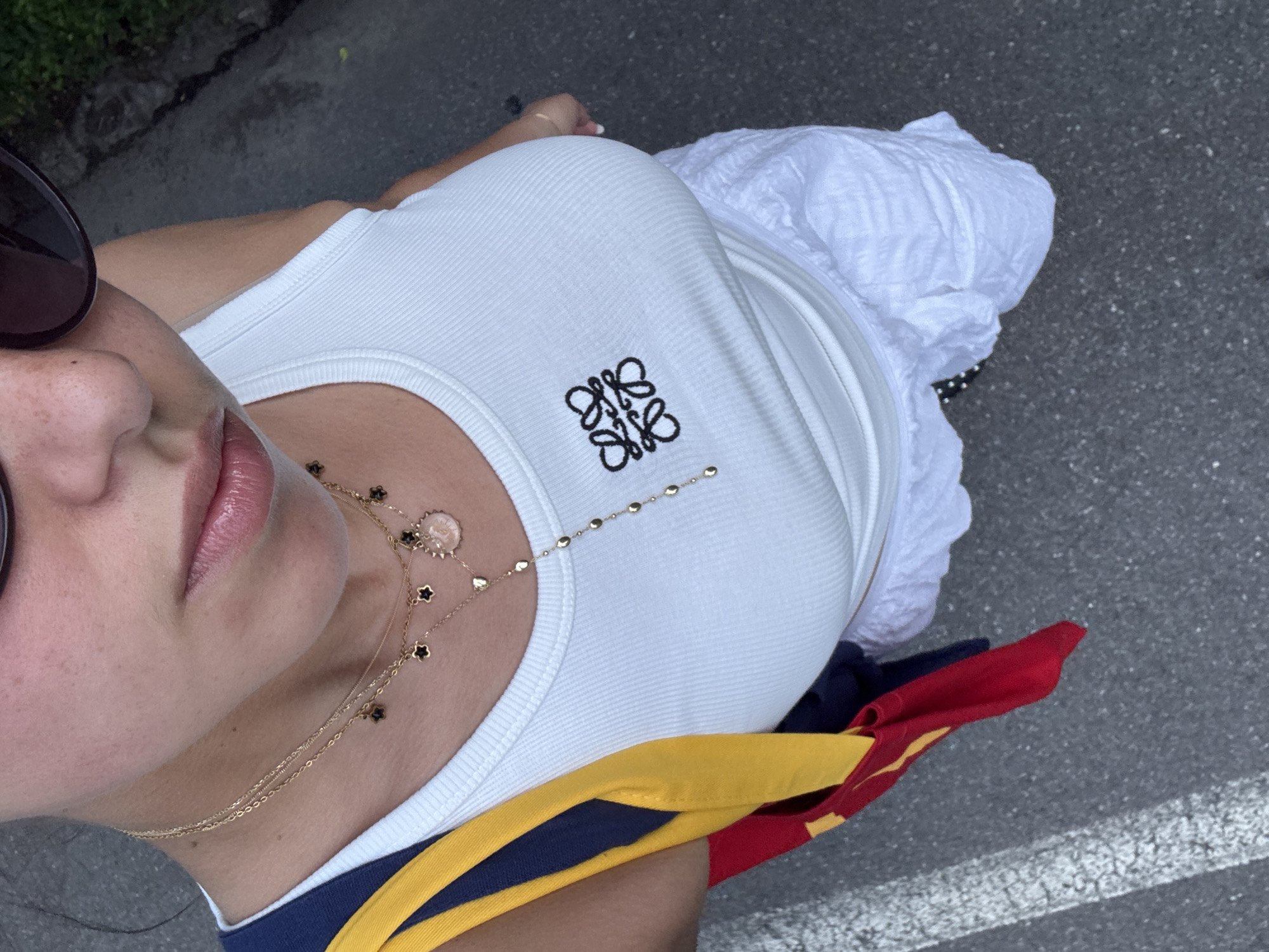 A high-angle, eye-level shot captures a person standing on asphalt pavement, presumed to be in Milan, Italy. The individual, appearing to be female, is observed from the chest down, with the upper forehead and hair cropped from the frame. She wears dark, oval-shaped sunglasses that obscure her eyes, and exhibits fair skin with visible freckles on the nose and cheeks. Her full lips are adorned with a light pink gloss or tint.
She is dressed in a fitted white ribbed tank top featuring a black embroidered Loewe Anagram logo on the left chest. Multiple gold necklaces are layered around her neck: two choker-style necklaces with small black four-leaf clover charms, a longer gold necklace with a circular pendant (possibly a coin or cameo), and a lariat-style gold chain featuring small disc-shaped beads extending vertically down the center of her chest. A thin gold bracelet is visible on her left wrist.
Her lower attire consists of a textured or ruched white skirt. Her left hand, with light-colored fingernails, is lightly grasping the hem of her skirt on the right side of the frame. A colorful strap or bag is slung over her right shoulder (viewer's left), displaying prominent bands of yellow, dark blue, and red fabric, with the red portion forming a pouch or bag hanging lower.
The ground beneath her is dark gray asphalt, showing signs of wear and a partial white painted line on the far left. The background is largely out of focus, revealing darker pavement and indistinct dark foliage in the upper right corner. The scene is brightly illuminated by natural light.
