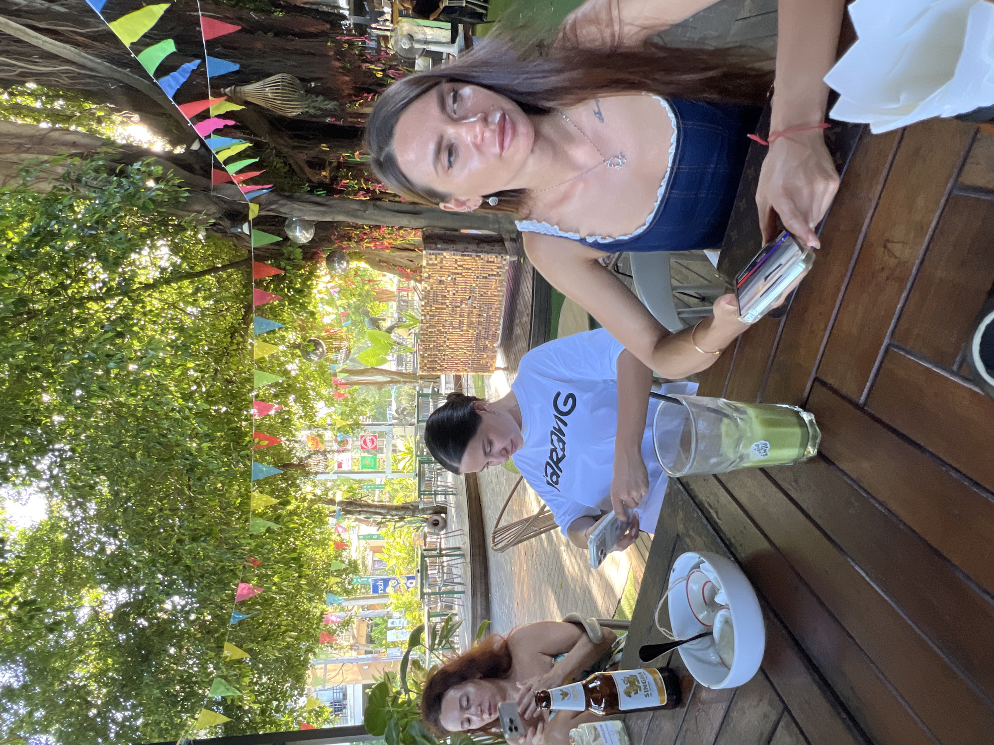 Three women are seated at a wooden table outdoors, possibly at a restaurant or cafe in Ban Bang Rak, Thailand. The woman on the far left has red hair and is holding a smartphone. The woman in the middle has dark hair pulled back and is wearing a white t-shirt with a black logo. She is also looking at a smartphone. The woman on the right has long dark hair and is wearing a blue denim top with white trim. She is holding a smartphone in her hands. On the table in front of them are a bottle of Singha beer, a bowl with food and utensils, and a glass of a green beverage. Colorful triangular flags are strung across the scene overhead, and lush green foliage and trees are visible in the background, suggesting an outdoor, tropical setting.