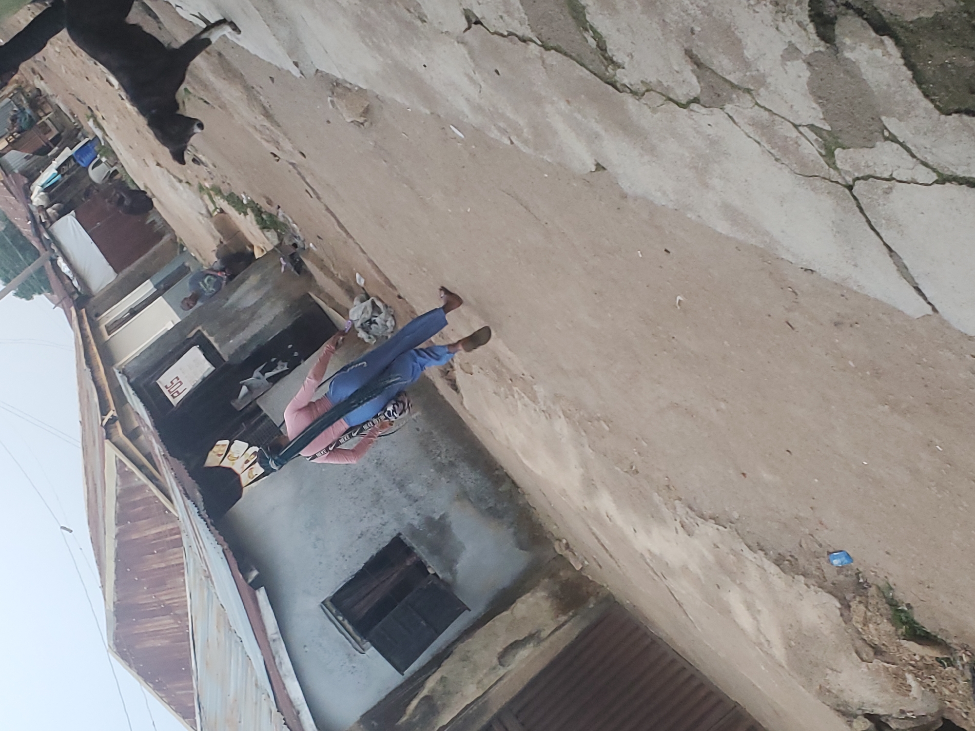 The image depicts an outdoor scene in Jos, Nigeria, featuring a person standing on what appears to be a cracked concrete or paved surface in front of buildings. The person is wearing a pink shirt and blue pants, and black Nike head wear. A black dog is visible in the upper left corner of the image. The buildings appear to be modest in construction, some with corrugated metal roofing. Other objects visible include building material and a cracked surface.