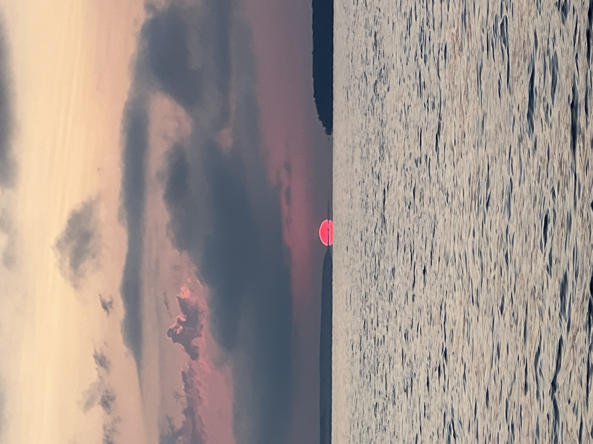 The image depicts a sunset over a body of water. The sun, a vibrant pink disk, is partially submerged on the horizon, casting a pinkish hue across the sky and clouds. Dark, textured clouds are scattered across the sky, with some illuminated by the setting sun. The water in the foreground is calm, with gentle ripples reflecting the colors of the sky. Silhouetted land masses are visible on the horizon to the left and right of the sun. The overall scene suggests a serene natural landscape. The image does not contain any people, cityscapes, or specific landmarks that would allow for identification of the location as Novopichugovo, Russia.