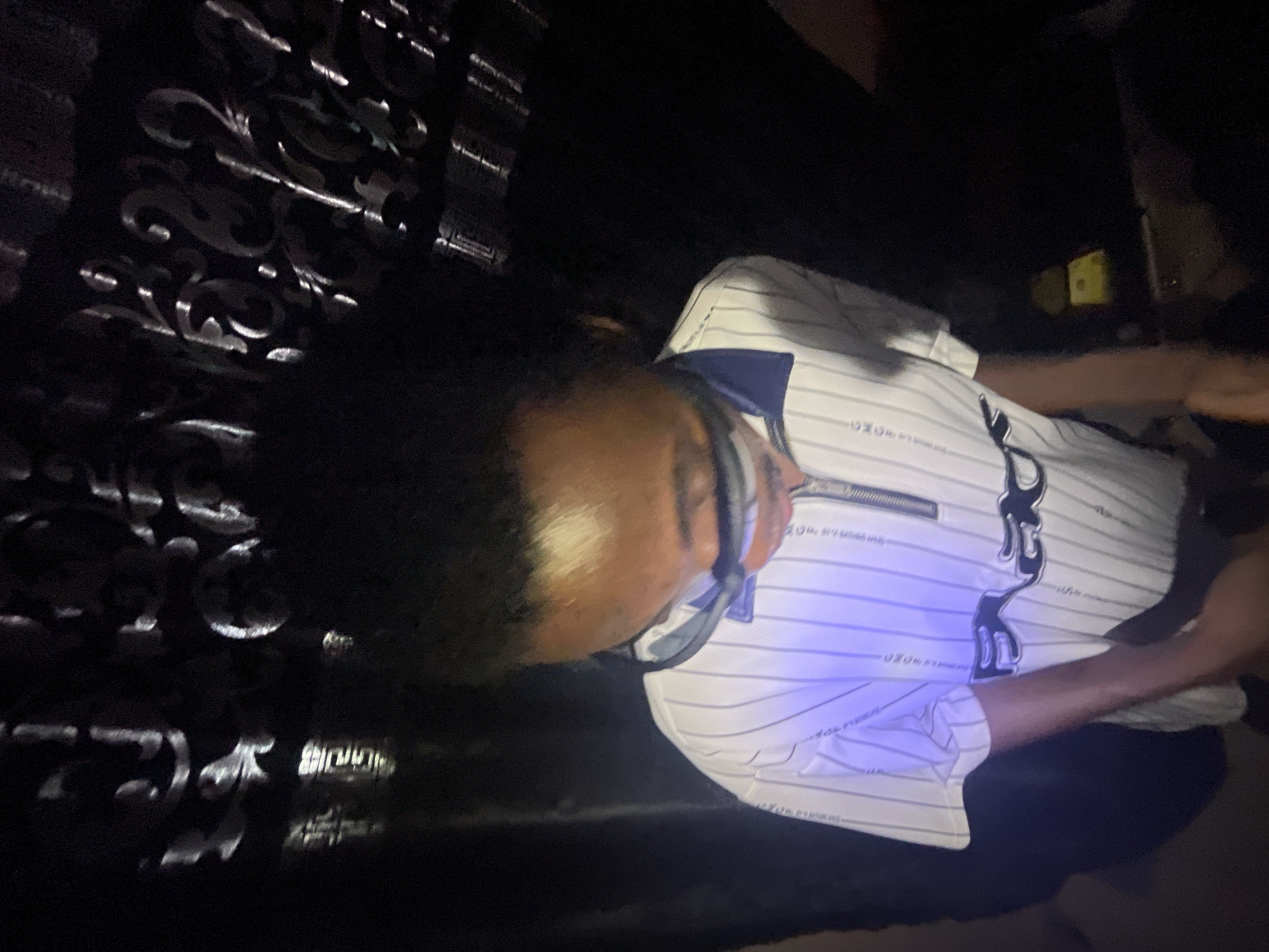 An individual is depicted indoors in what appears to be a dimly lit setting. The person has dark curly hair and is wearing glasses. They are dressed in a short-sleeved, striped shirt with a navy blue collar and distinctive designs on the front. The background features ornate metallic or reflective patterns, possibly a gate or screen. The scene is dark, making it difficult to discern specific details. No explicit information confirms the image location in Lokoja, Nigeria.