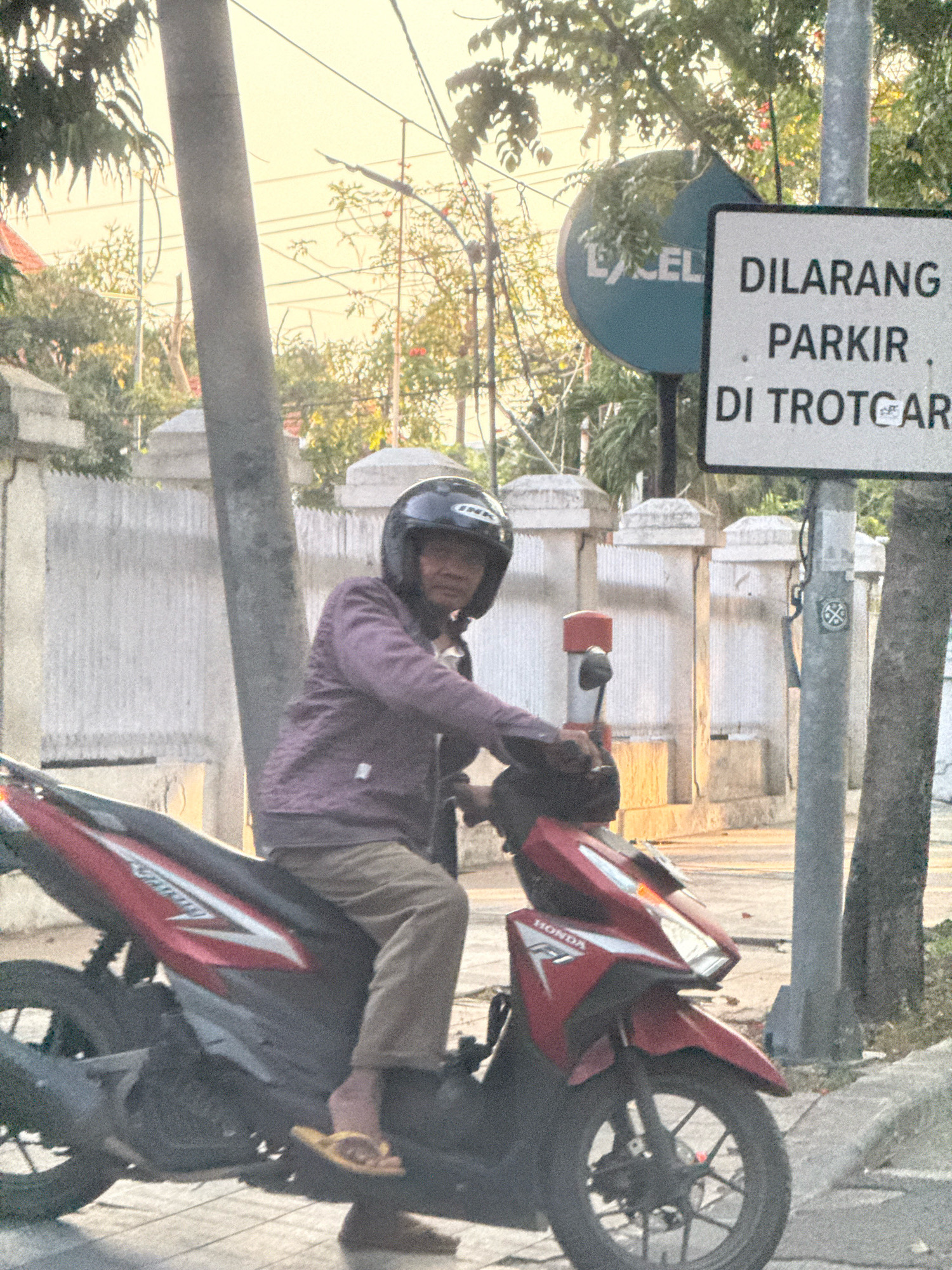 The image shows a person riding a red Honda motorcycle on a street in Surabaya, Indonesia. The rider is wearing a dark purple jacket, light brown pants, and a black helmet. The motorcycle has "HONDA" and "F1" branding visible on its front panel. A white and black sign is posted on a pole to the right of the motorcycle, which reads "DILARANG PARKIR DI TROTOAR," meaning "No Parking on the Sidewalk" in Indonesian. A circular blue sign with white text is visible in the background, partially obscured by trees, which appears to say "EXCEL". The environment is an urban street scene with a sidewalk, a paved road, and some vegetation. The lighting suggests it might be daytime, possibly late afternoon, given the warm tones.