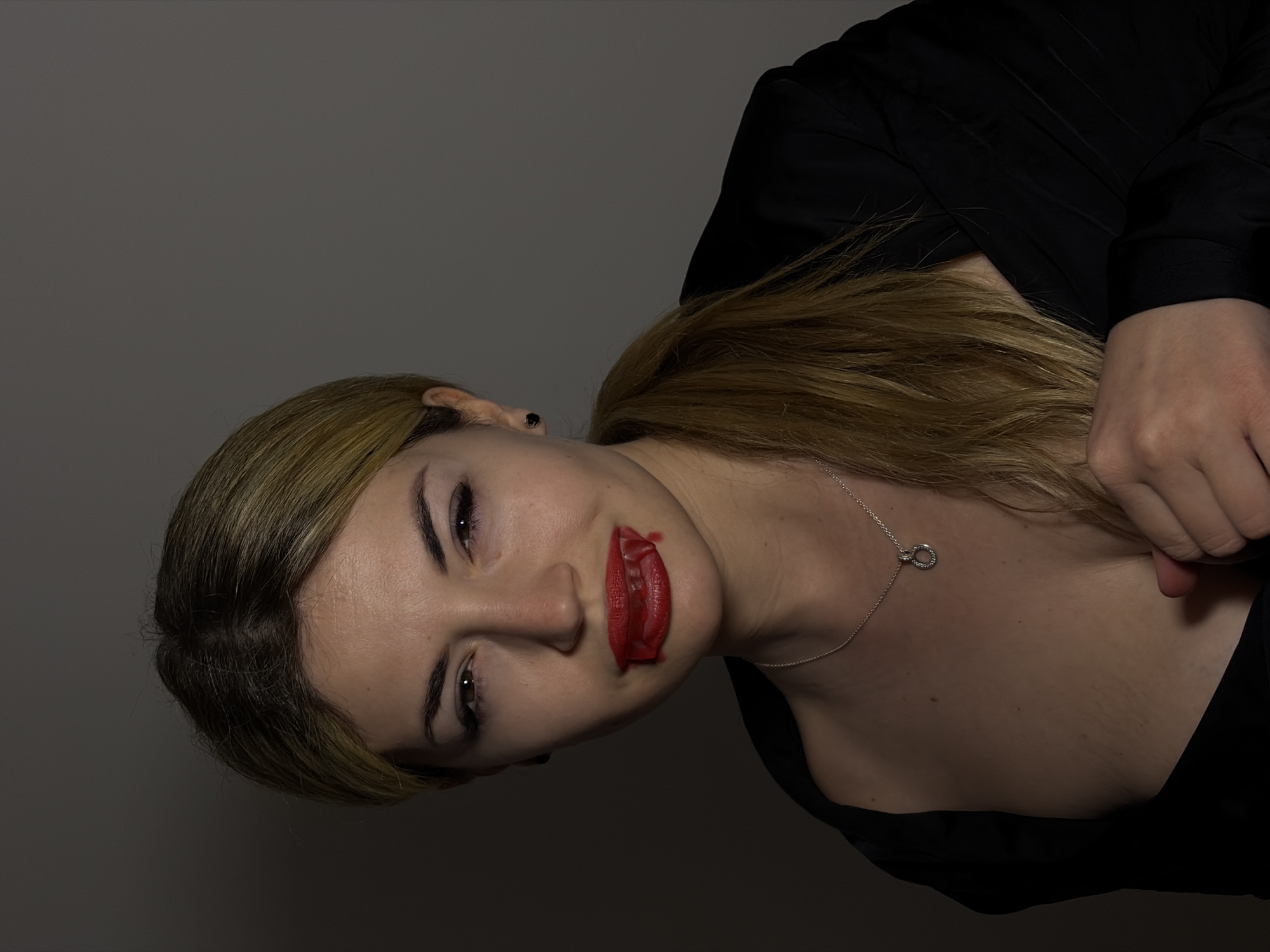 The image shows a woman with blonde hair and red lipstick smeared around her mouth. She is wearing a black top and a silver necklace with a circular pendant. The background is a neutral gray. The image does not contain information about the city of Budva, Montenegro.