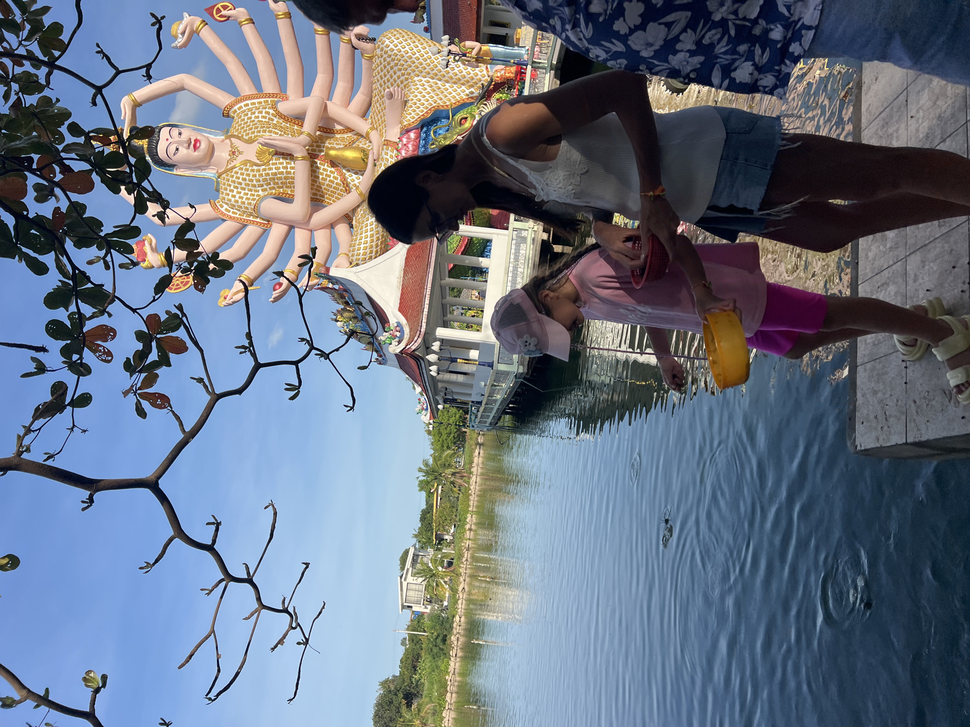 The image shows a scene in Baan Plai Laem, Thailand. A large, golden statue of a deity with multiple arms stands in the background, partially obscured by tree branches. An adult and a child are positioned at the edge of a body of water, likely a lake or pond. The adult is wearing a white top and denim shorts. The child is dressed in a pink shirt and pink shorts, wearing a baseball cap. The child is holding a yellow container and appears to be interacting with the water. Reflections of the buildings and statue are visible in the water. Ripples in the water indicate possible movement or objects submerged. Another person is partially visible in the upper right corner. The background features buildings and greenery, indicating an urban or semi-urban environment. The sky is clear and blue.