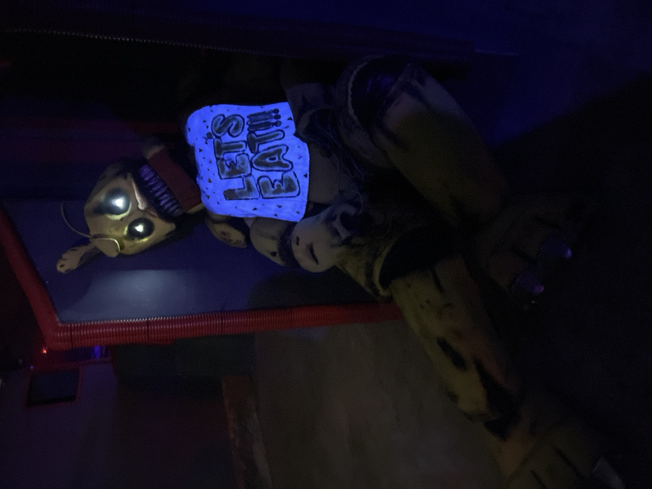 The image displays a dark, low-light scene featuring a horror-themed animatronic or character figure lying horizontally across the frame. The overall atmosphere is dim, with blue and subtle yellow light sources illuminating parts of the subject.
The central figure is a worn, yellowish-green character, resembling a damaged animatronic. Its head, positioned towards the left side of the frame, features two glowing white or pale yellow circular eyes that stand out against the darker head material. Below the eyes, a mouth-like area shows exposed, jagged, or teeth-like structures, possibly indicating damage or a menacing design. Small wires or mechanical components are visible around the head and neck area.
The most prominent feature is a bright, glowing blue rectangular area on the character's chest. This area contains white text, also glowing, that reads "LET'S EAT!!!" in a stylized, slightly uneven font with three exclamation marks. The blue glow from the chest illuminates the immediate surroundings.
The body of the character appears to be made of a textured material, possibly fabric or plastic, that is distressed and dirty, with darker smudges and patches suggesting age or damage. Visible arms extend from the torso, and one hand is partially visible on the right side of the frame. The fingers of this hand have distinct purple tips, which could be painted nails or part of the character's design.
In the lower left portion of the image, beneath the character's head, there is a distinct red, corrugated or ribbed structure, possibly a pipe, a decorative element, or part of a bed frame or platform. The background is largely indistinct and dark, contributing to the mysterious and eerie mood of the image. The character appears to be lying on a dark, flat surface.
The image is captured in a dark environment, possibly indoors at night or in a dimly lit room, with specific lighting used to highlight the character and its glowing features. The blue light suggests the use of blacklight or UV light.