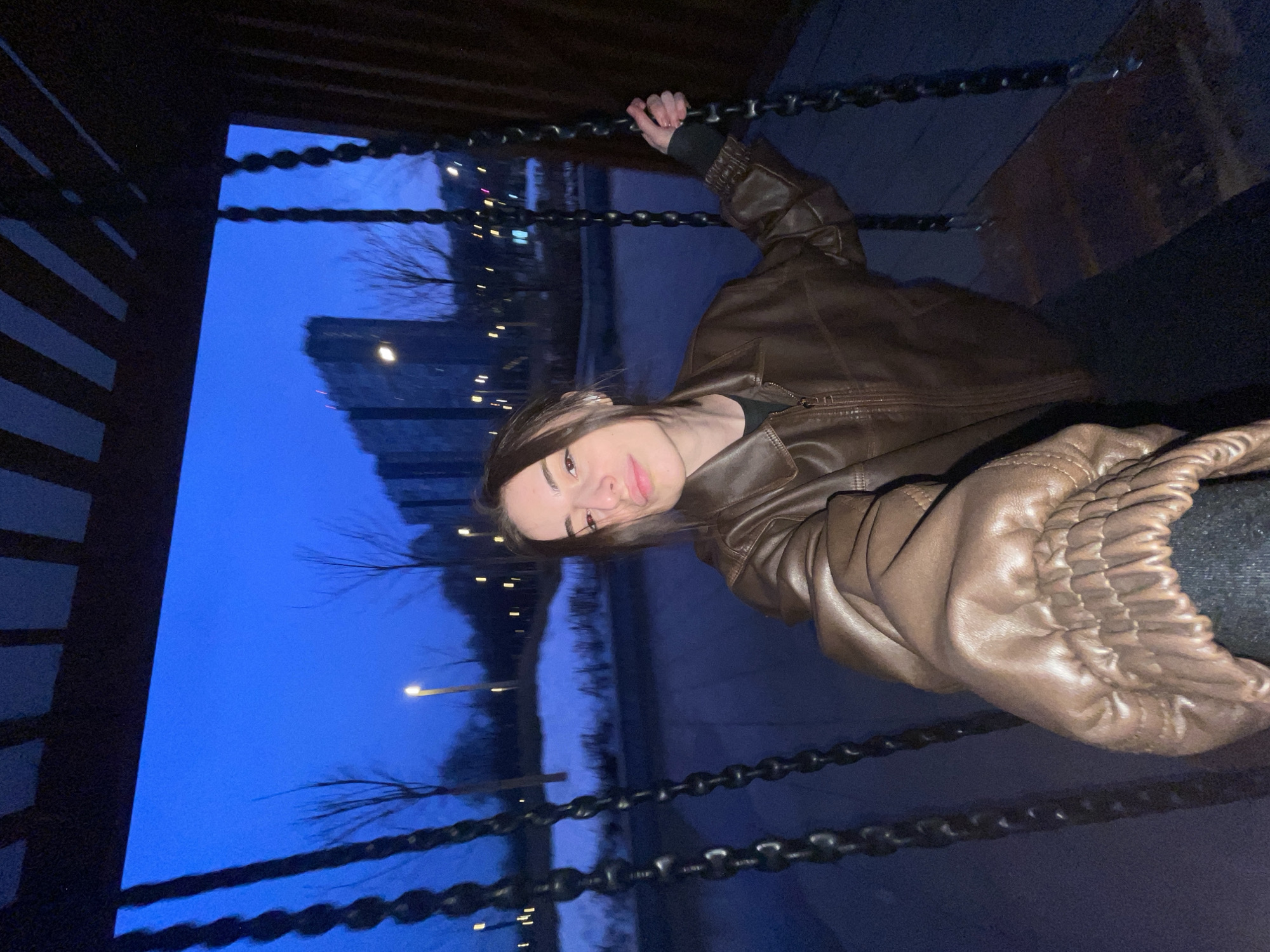 A female individual is seated on a swing within Khodynskoye Pole Park in Moscow, Russia, at twilight or night. She wears a brown leather-like jacket over a dark shirt and dark trousers, and holds the swing chain with her left hand while extending her right arm towards the camera for a selfie. Her gaze is directed at the camera with a slight smile. The swing structure, featuring a dark, slatted canopy, frames the upper portion of the scene. In the background, patches of snow cover the ground, surrounded by bare deciduous trees and illuminated by scattered streetlights. Beyond the park, several modern, high-rise buildings, consistent with the ZILART residential complex, are visible against the deep blue sky.