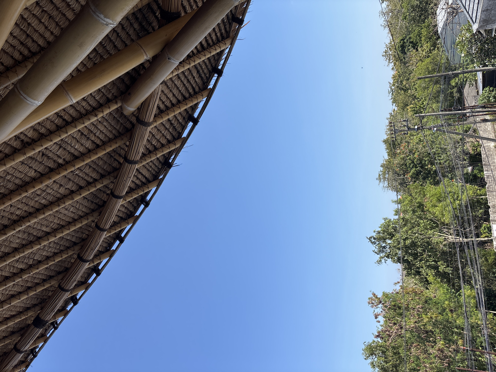 The image captures an outdoor scene in Kuta Selatan, Indonesia, viewed from a low angle. The upper portion of the frame is dominated by the underside of a thatched roof structure, extending diagonally from the top-left to the top-right. This roof is constructed from densely woven, dark brown natural fibers, likely palm leaves or similar thatch, supported by a framework of light brown bamboo poles. Larger, cylindrical bamboo beams are visible, with dark bindings securing them.
Below the roof and occupying the majority of the mid-frame is a clear, cloudless, light blue sky.
The lower third of the image reveals a distant treeline composed of various green trees, some with brown foliage, indicating a lush landscape. Beneath the treeline, an urban or semi-urban environment is present, featuring multiple black power lines strung horizontally across the frame. These lines are supported by several utility poles, visible as dark vertical structures. In the bottom-right corner, parts of a light grey concrete wall and fragmented views of building rooftops can be discerned. There are no individuals or visible actions depicted in the scene.