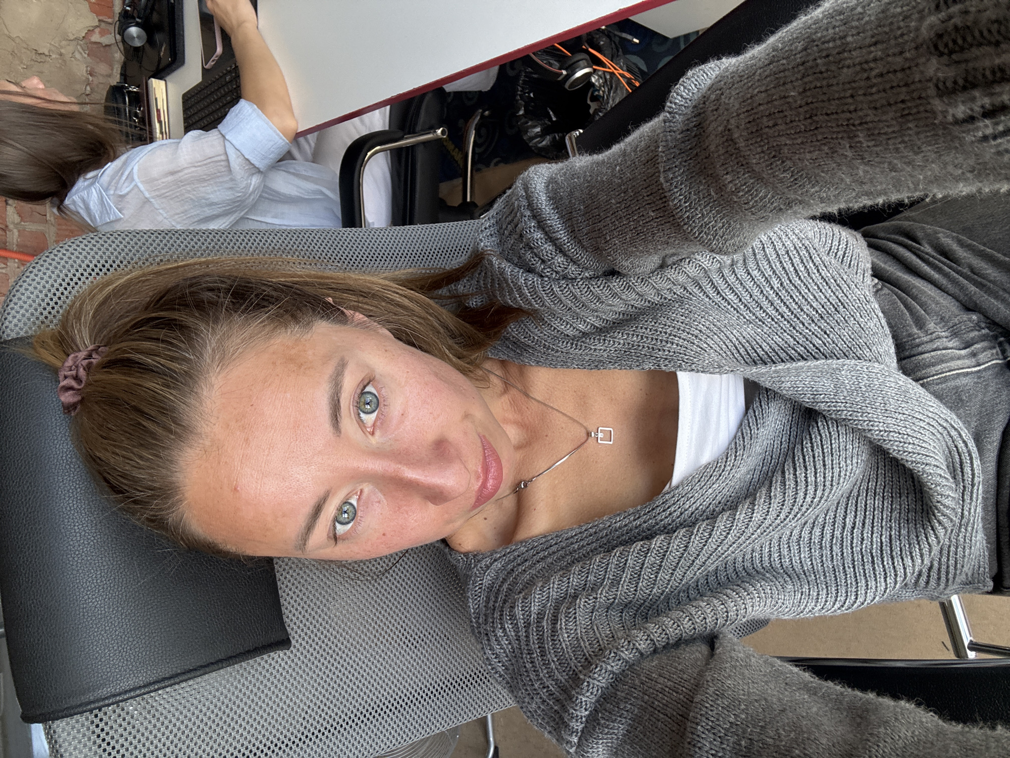 This is a selfie taken from a high angle, looking down at a woman lying on her back. She has light brown hair pulled back with a scrunchie and light green eyes. She is wearing a grey knit cardigan over a white top and a silver necklace with a small square pendant. The woman appears to be in a room with a desk and a chair. A second person, partially visible, is sitting at the desk wearing a light blue collared shirt. The room has a brick wall in the background. The overall lighting suggests it is daytime.