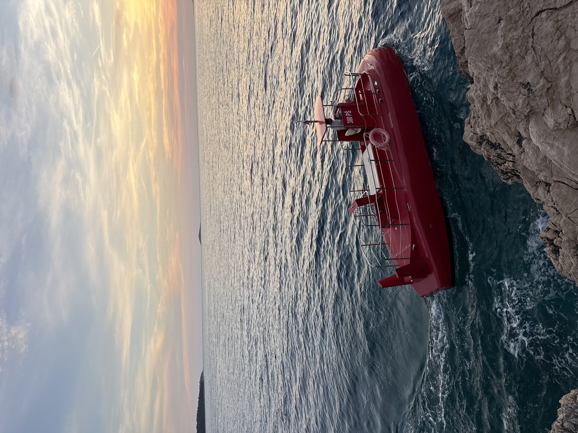 A red submarine-like vessel with a canopy is seen in the turquoise waters of the Adriatic Sea near Primošten, Croatia. The vessel is observed from a rocky shoreline, with a person visible in the pilot's seat. The sky above is a gradient of pale blue and orange, indicating either sunrise or sunset, with some wispy clouds. The sea has gentle waves, and a small island is visible in the distance.