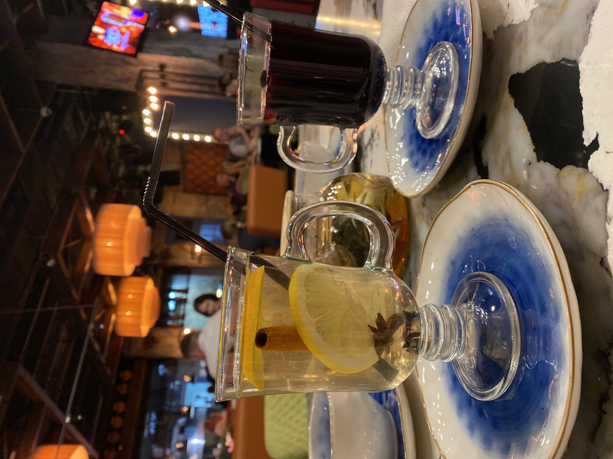 The image shows two drinks, one dark red and one light yellow, both served in glass mugs with straws, placed on blue and white saucers. The light yellow drink contains lemon slices, a cinnamon stick, and star anise. They are positioned on a marbled surface. In the background, there are blurred lights, furniture, and people, indicating an indoor setting, likely a restaurant or cafe. A television screen is visible in the upper left. The location is Yekaterinburg, Russia, as the image was sourced from there.