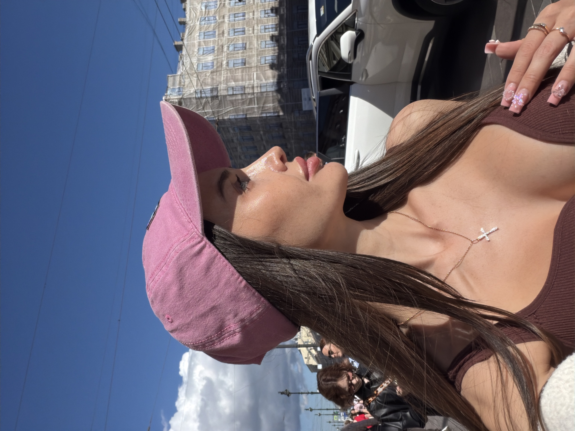 In this medium shot, a young woman is depicted outdoors on a sunny day. She is wearing a pink baseball cap and a brown tank top. A delicate silver cross necklace is visible around her neck. Her long, dark hair is down and falls over her shoulder. Her fingernails are long, square-shaped, and adorned with intricate pink glittery designs and small rhinestones, and she wears several rings on her fingers.
In the background, a building under construction, covered in white scaffolding, is visible against a bright blue sky. Power lines crisscross the sky. Other people are blurred in the background, suggesting a public place like a street or plaza. The lighting indicates it is daytime, likely midday given the bright sun. The overall scene is casual and summery. The image has been rotated by 90 degrees clockwise.