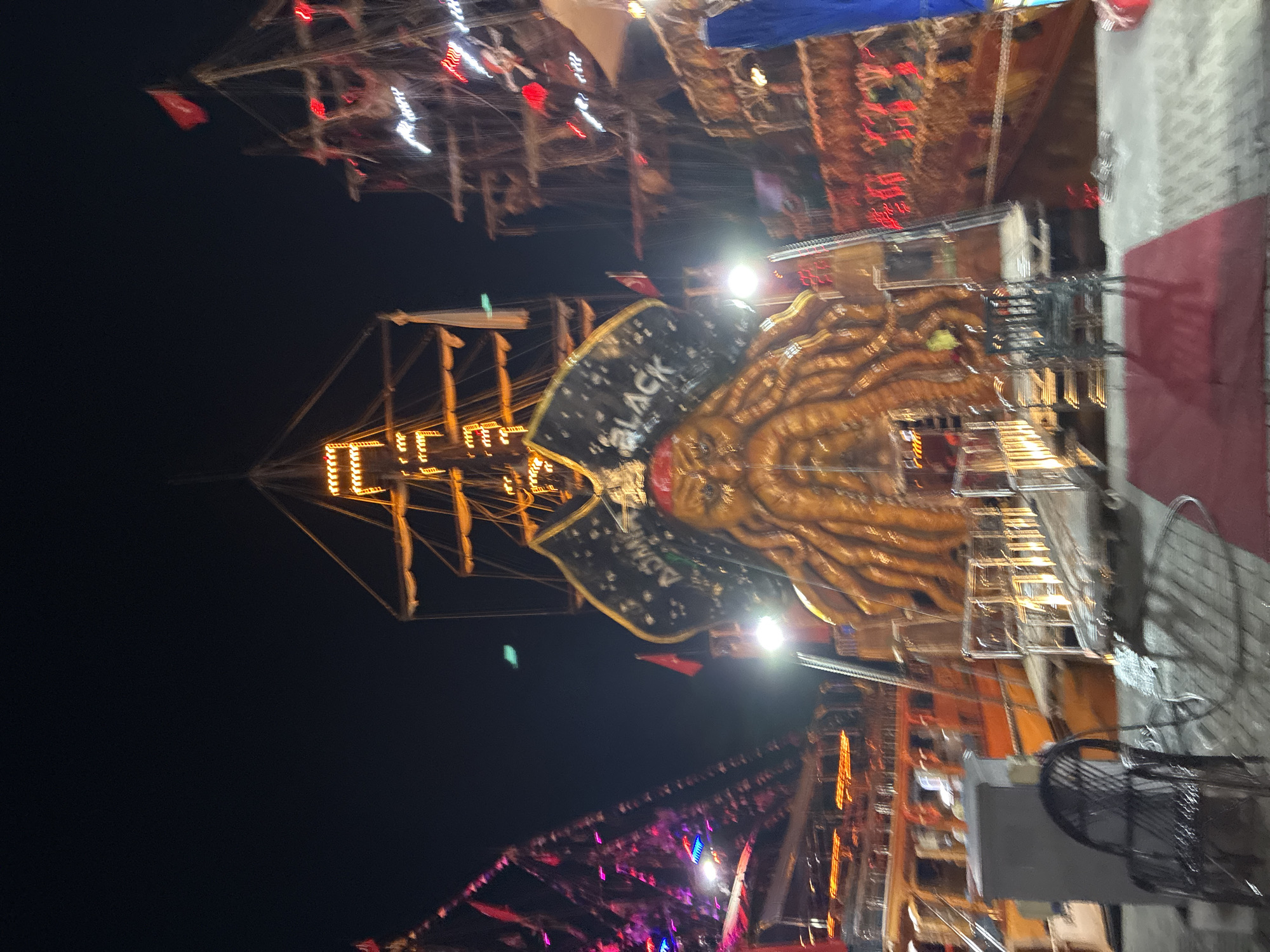 The image depicts a night scene at a harbor in Alanya, Turkey, likely a tourist attraction. Several large, elaborately decorated pirate-themed ships are moored together. The central ship features a prominent, large, golden octopus sculpture on its bow, with a pirate flag behind it. The ships are illuminated with numerous colorful lights, including strings of yellow and red bulbs along the masts and hulls, as well as pink and blue lights on the ship to the left. The overall atmosphere is festive and somewhat chaotic due to the motion blur, suggesting the photograph was taken with a slow shutter speed or during movement.
In the foreground, there is a paved walkway with red mats laid out. A dark-colored metal chair is visible on the left, and part of a railing system is present. The background is dominated by the dark night sky, with the lights of the ships providing the primary illumination. The image appears to be taken from a vantage point on the dock or a nearby vessel. The blurred nature of the image suggests it might be a nighttime tourist spot that comes alive with lights and entertainment.