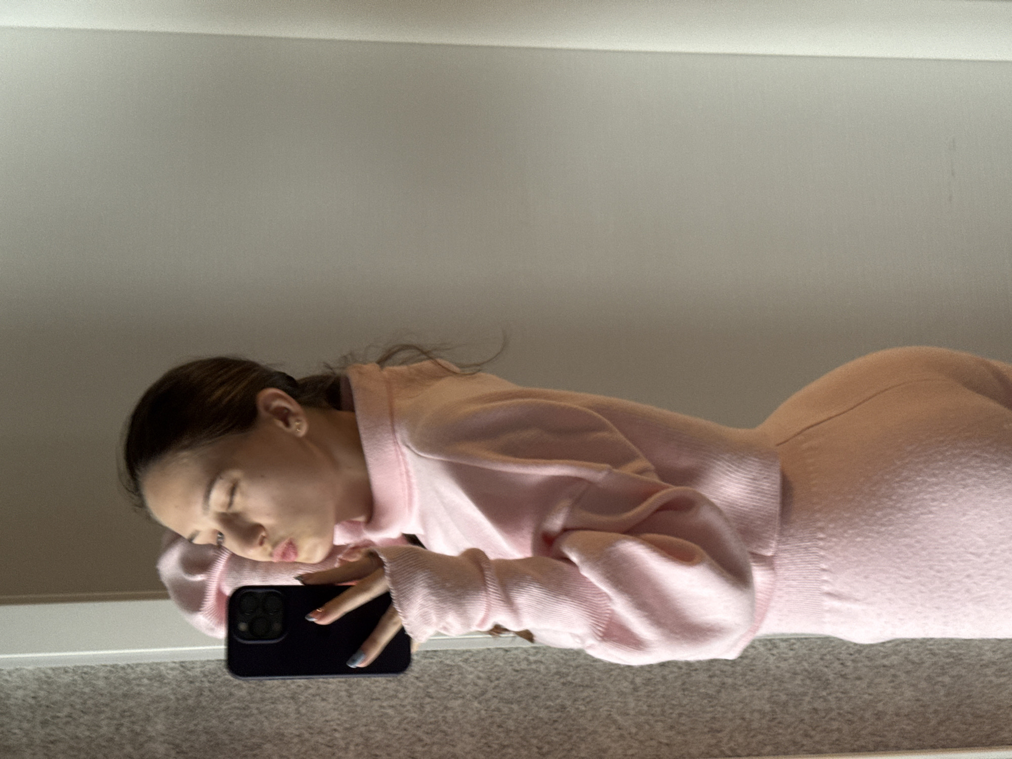 A woman is lying on her side on a gray carpet, with her head resting on a black smartphone. She is wearing a pink sweater and what appears to be a matching skirt. The background is a plain white wall. There is no evidence to determine the location in the city of Sofia, Bulgaria.