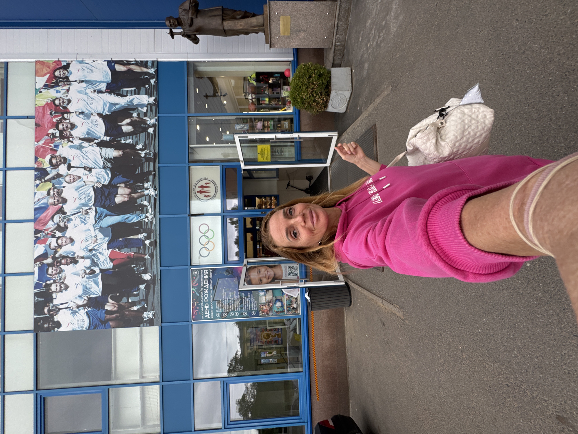 The image is an eye-level shot of a blonde woman in a pink sweatshirt, holding the camera in her right hand. She is standing in front of a blue and white building, likely in Saint Petersburg, Russia, as Cyrillic text is visible.
The building features a large mural or advertisement of people running with flags, possibly related to a world run event. The Olympic rings are also visible on the building, suggesting it is related to sports or athletics. There is a sign advertising birthday parties ("День Рождения"), with an offer for 2000 Rubles per hour on the LOFT venue ("АКЦИЯ 2000Р/ЧАС НА АРЕНДУ ЛΟΦΤΑ"). A statue stands on a pedestal above the building's entrance.
The environment appears to be urban, with a gray paved area where the woman is standing. A white bag is on her left.