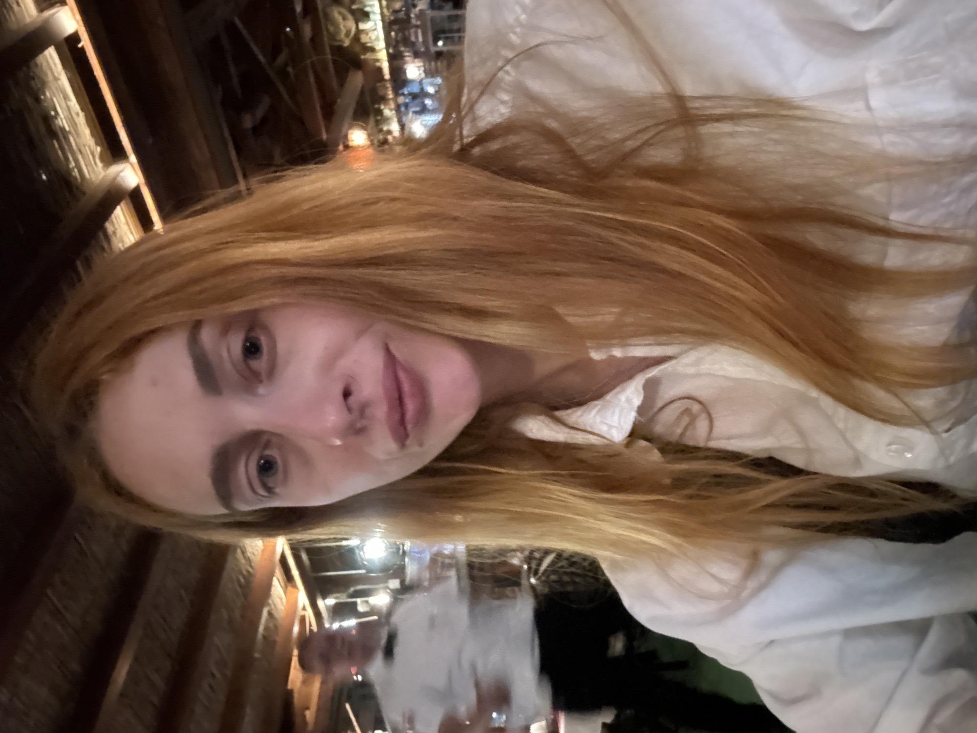 A young, fair-skinned woman with long, wavy, strawberry blonde hair is seen indoors, likely at a restaurant or bar, based on the wooden architecture and lighting visible in the background. She is wearing a white collared shirt. The scene includes indistinct figures in the background. While it is not possible to pinpoint the exact location within Budva from this image alone, the architectural style suggests a venue that is consistent with coastal regions. There are no visible actions, and the subject is looking directly at the camera, possibly taking a selfie.