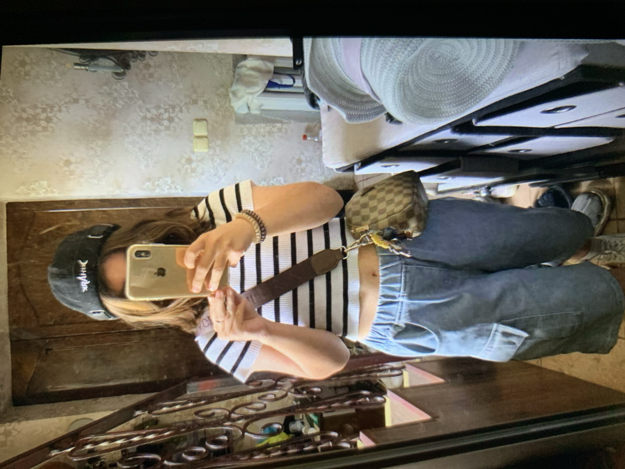 A person is taking a mirror selfie in what appears to be a residential interior. The person is wearing a black and white striped crop top, light blue cargo shorts, a black baseball cap, and sneakers. They are holding a gold smartphone in their right hand to take the photo. A brown crossbody bag with a checkered pattern is slung across their body. The background includes a wooden staircase, a door, wallpaper with a floral pattern, light switches, a bottle of liquid, and a stack of drawers with hats on top. The image does not contain any information that can be used to determine the location in the city of Lymanka, Ukraine.