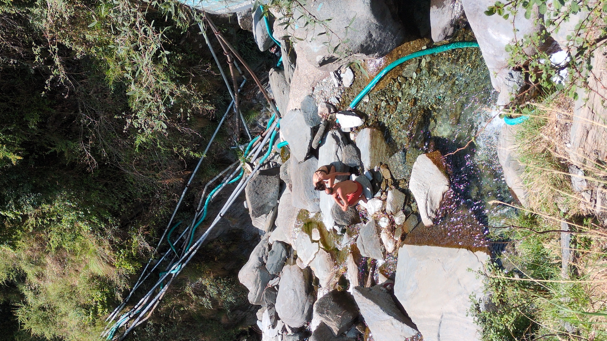 The image captures a scenic view in Bhagsu, India, featuring a rocky stream. Two individuals, a woman in a bikini and a man in red shorts, are seated among the rocks near the water. A blue hose runs alongside the stream, seemingly supplying water. The environment is lush with greenery on the surrounding banks. Metal supports are visible amongst the vegetation, possibly for infrastructure related to water management or crossing. The scene gives the impression of a natural spring or stream being utilized for recreational purposes amidst a natural setting.