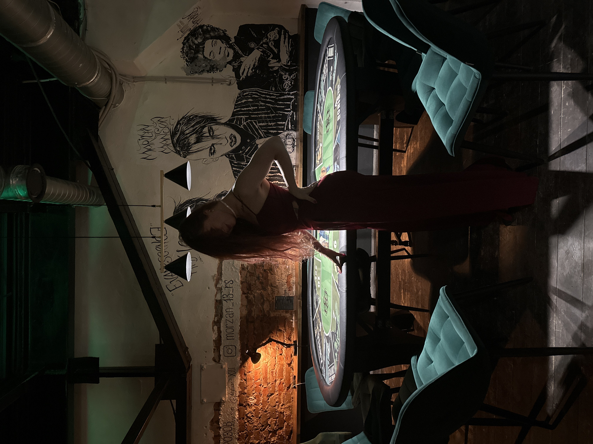 The image shows the interior of a dimly lit room, appearing to be a bar or club, possibly located in Vladimir, Russia, based on the writing on the wall. A woman in a red dress is leaning over a round poker table with a green surface, touching the table with her right hand. Around the table are several teal colored upholstered chairs. On the wall behind her are black and white drawings of people, including one of Jim Morrison, and graffiti style writing. Above, there are industrial-style ductwork and hanging pendant lights. The room appears to have exposed brick and structural beams. The lighting is dark and uneven, with some areas more illuminated than others.