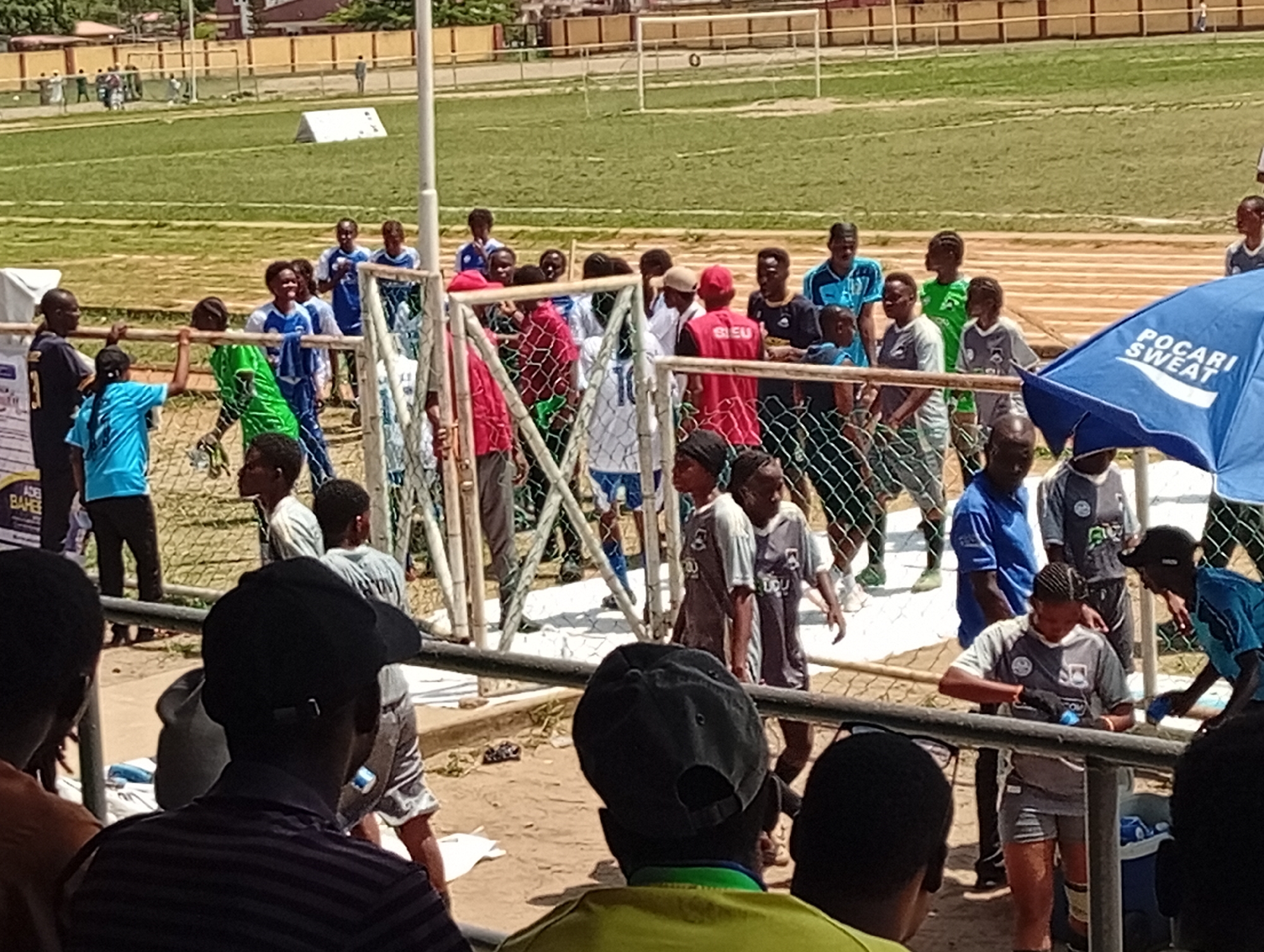The photograph shows a soccer game taking place at a stadium in Lagos, Nigeria. The stadium has a green soccer field with white boundary lines and goalposts. In the background, a low, beige building runs along the edge of the stadium.
A group of people, including players in soccer uniforms, are gathered near a fence that separates the field from the stands. Some players are wearing blue and white uniforms, while others are wearing gray and black. A person in a green jersey is also visible. A blue "POCARI SWEAT" umbrella is positioned near the fence, likely for shade.
In the foreground, the backs of the heads of several spectators are visible, suggesting they are watching the game. A sign with the text "ADE BAHE" is visible on the left side of the image. The photograph appears to have been taken from the stands, looking out over the soccer field. The time of day seems to be daytime, with sunny conditions.