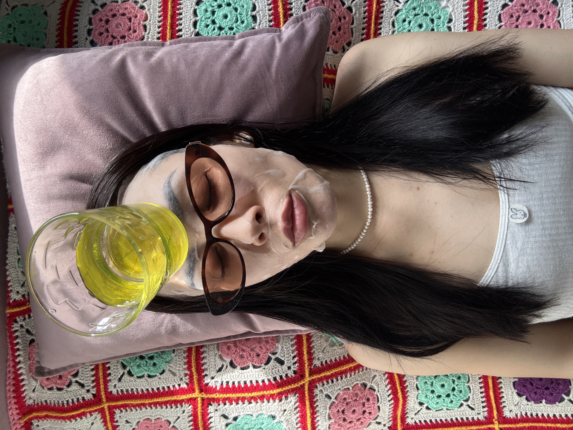 A woman is lying on a multicolored crocheted blanket. Her head rests on a plum-colored pillow. She is wearing a face mask and sunglasses, and a glass of yellow liquid is precariously balanced on the pillow near her head. She has long, dark hair and is wearing a pearl necklace and a white tank top with a bunny logo. The location is not discernible as Votkinsk, Russia, from the information provided in the image.