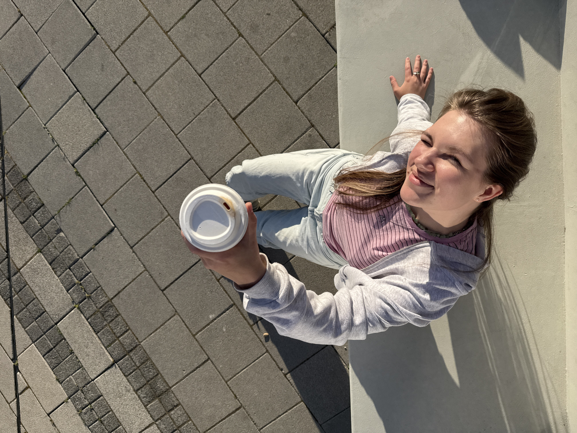 A young woman stands outdoors in Belgrade, Serbia, holding a coffee cup with a white lid in her left hand. She is positioned adjacent to a light gray concrete wall. The ground beneath her feet is paved with gray rectangular stone blocks. The woman is wearing light blue jeans, a pink ribbed top, and a gray zip-up hoodie. Her hair is pulled back. She is looking upwards and smiling slightly. Sunlight casts shadows on the wall next to her. Her right hand rests on the concrete wall. A ring adorns her right hand.