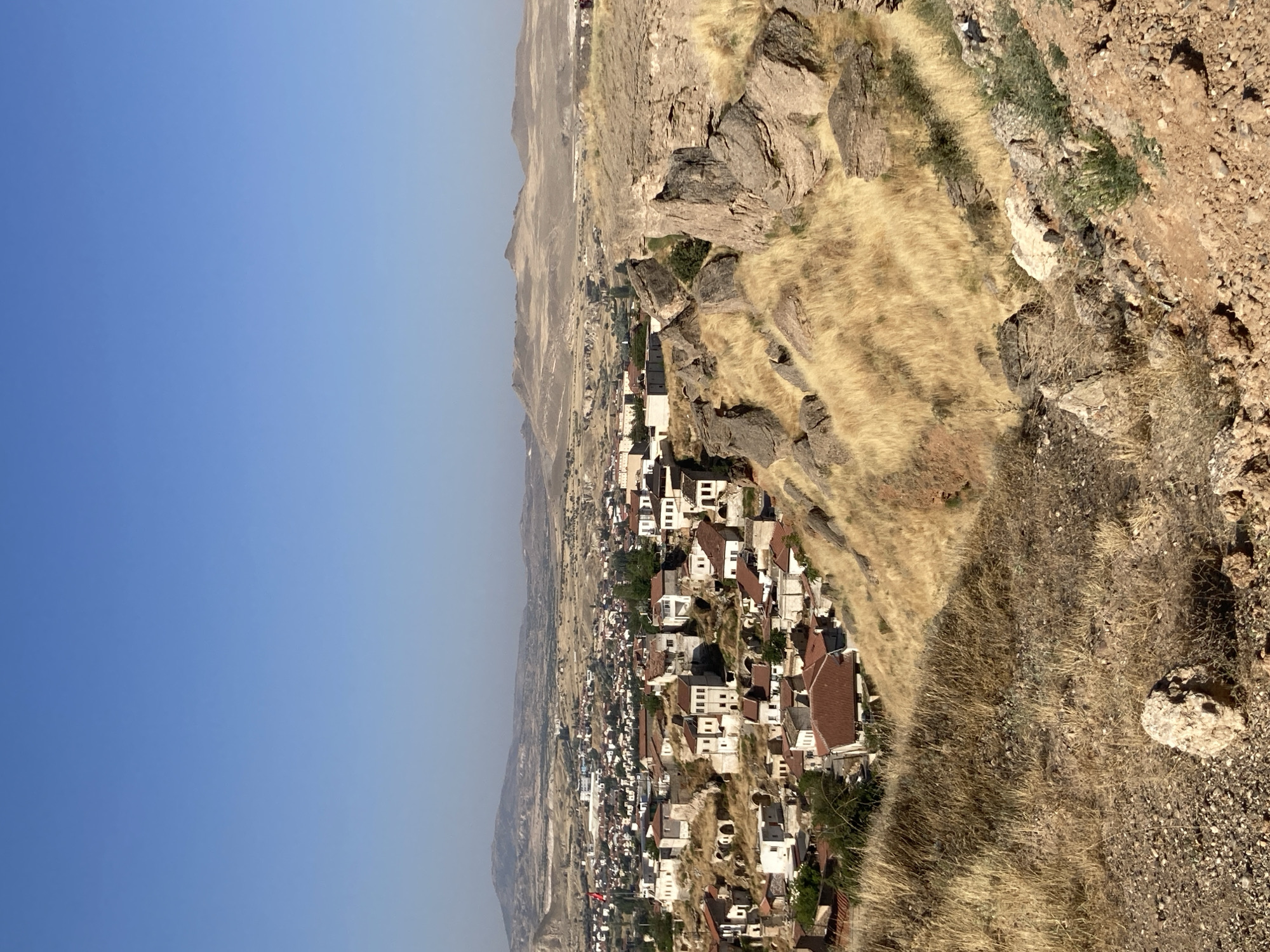 The image captures a panoramic view of Avanos İlçe Merkezi, Turkey, on a clear, sunny day. In the foreground, a rocky, dry, and sparsely vegetated hillside slopes downwards. This hillside is covered with dry, yellow grass and scattered rocks of various sizes, with a larger, lighter-colored rock prominently positioned in the lower left quadrant.
Further down the slope, the landscape transitions into a densely populated urban area. Numerous buildings, predominantly with red-tiled roofs and light-colored walls, are clustered together. Many of these structures appear to be built into the natural rock formations, showcasing a distinctive architectural style. Some buildings have multiple stories, with visible windows.
In the middle ground, the town extends outwards, with more buildings and some scattered trees visible. A Turkish flag is mounted on a flagpole on the left side of the town.
The background of the image features a vast, arid landscape characterized by rolling hills and distant mountains. The terrain appears dry and rocky, with muted earthy tones. The sky above is a clear, cloudless blue, indicating fair weather conditions.
No people are visible in the image. The scene depicts a static, established settlement within a rugged, natural environment.