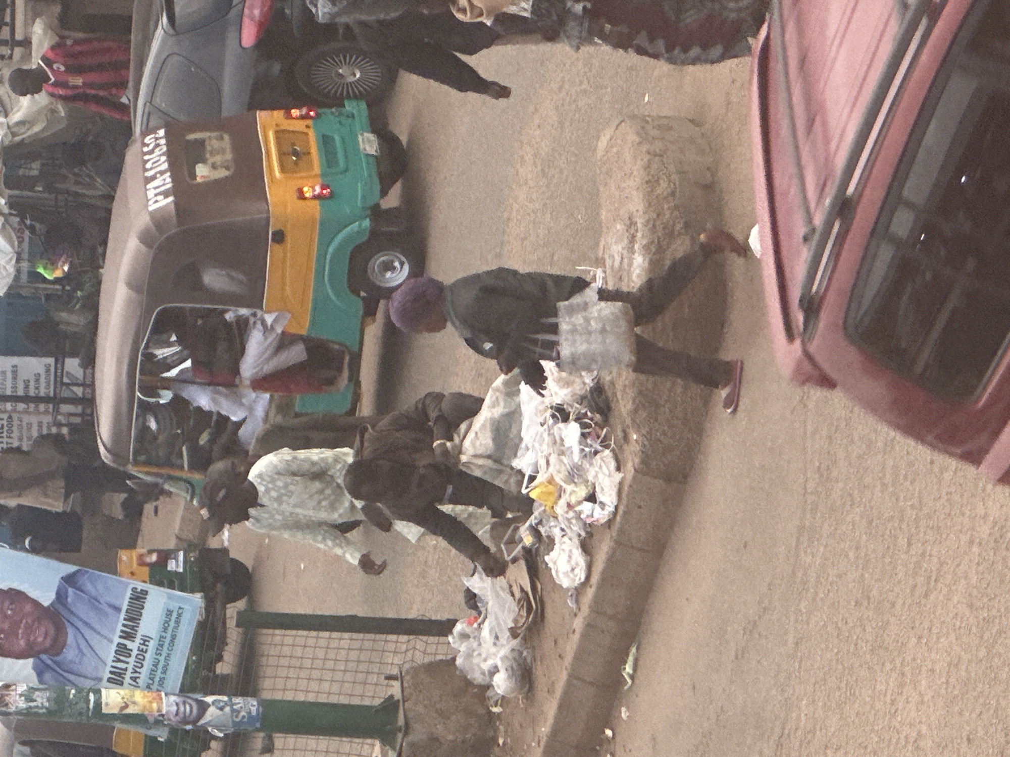 The image shows a street scene in Bukuru, Nigeria, with a focus on the activities around a small, three-wheeled vehicle, commonly known as a "Keke Napep." The Keke Napep, colored yellow and turquoise, is parked on the right side of the frame, its license plate reading "PTA-10652." A man in a white patterned shirt and dark pants is bent over, seemingly sorting through a pile of trash on the sidewalk. Nearby, another man in dark clothing is also engaged with the refuse. To the left, a prominent poster displays the face of a man identified as "DALYOP MANDUNG (AYUDEH)," with text indicating "PLATEAU STATE HOUSE" and "JOS SOUTH CONSTITUENCY." Another smaller poster featuring a smiling face is affixed to a green pole. In the background, additional vehicles and pedestrians are visible, contributing to the bustling atmosphere of the urban environment. A dark-colored car is also present in the scene, to the right of the Keke Napep. The overall impression is one of everyday life in a Nigerian city, with elements of commerce, waste management, and political messaging.