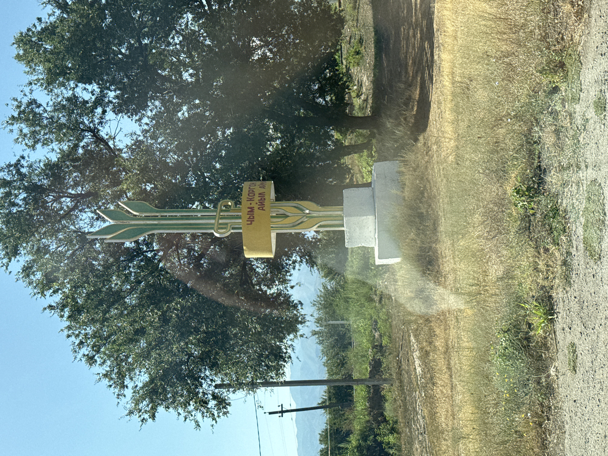 The image shows an outdoor scene in the city of Chim-Korgon, Kyrgyzstan. In the foreground, a white concrete monument stands in dry, yellowed grass. The monument is topped by a tall, stylized green and gold structure resembling wheat stalks. A yellow rectangular sign is attached to this structure with text that appears to be in Cyrillic script. Behind the monument, a large tree with dense green foliage is prominent. In the middle ground, a dirt path leads away from the monument, and more trees and vegetation are visible. Further in the background, a utility pole with wires stands against a backdrop of distant mountains under a clear blue sky. The overall impression is of a rural or semi-rural setting. There are no people or animals visible in the image.