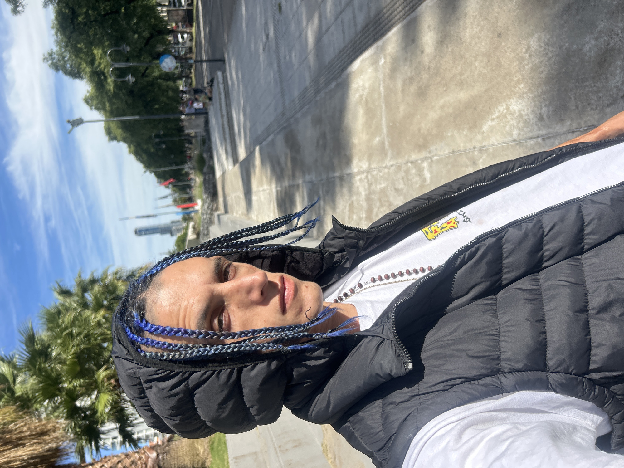 A medium shot of a man with dark hair braided with blue extensions. He is wearing a black puffer vest over a white T-shirt. A beaded necklace is visible. The background includes a cityscape, trees, and a paved area that appears to be a walkway or public space. The sky is blue with scattered clouds. Based on the architectural features and vegetation, the location is likely in Buenos Aires, Argentina.