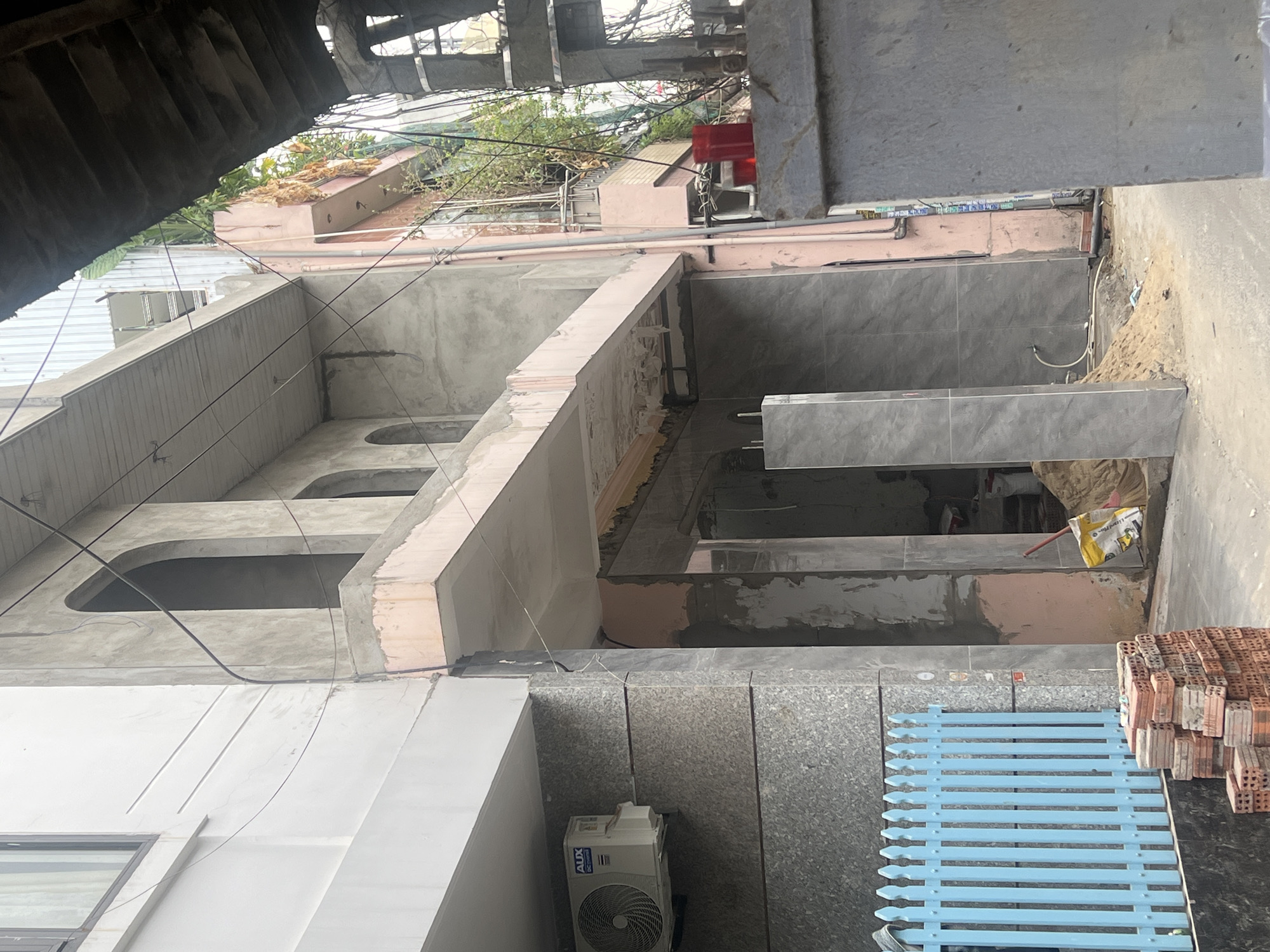 The image depicts a construction site in an urban setting, possibly in Đà Nẵng, Vietnam, given the architectural style and building materials. The foreground features stacks of red bricks and a blue, slatted metal structure. An air conditioning unit is mounted on the lower wall. The building is under construction, with exposed concrete and unfinished surfaces. Arched openings are visible on the facade. The upper levels of the building have gray tile work. Overhead, wires crisscross the view, indicating the urban environment. A red object is visible in the upper middleground. The overall scene is one of ongoing construction activity.