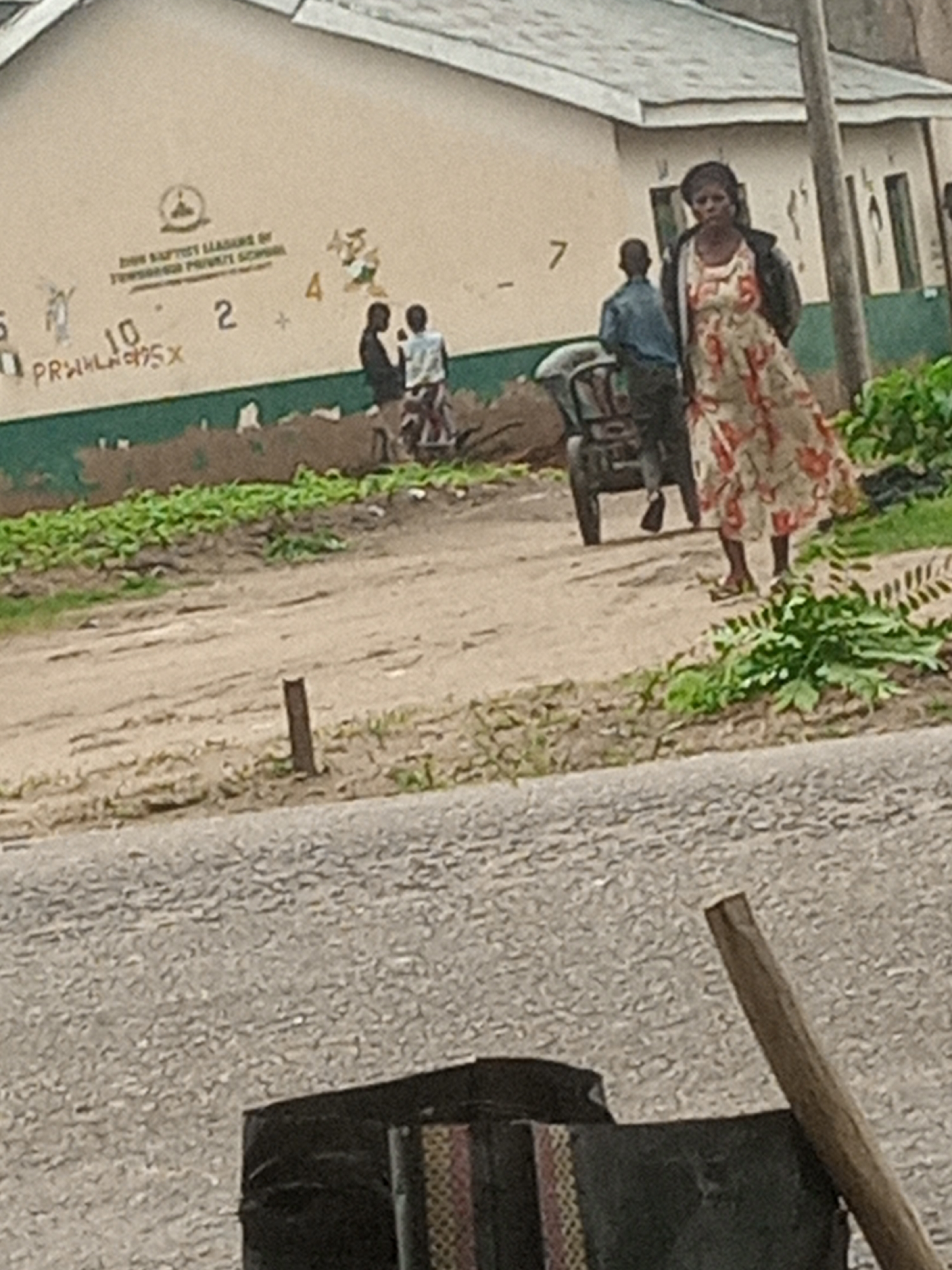 In Langtang, Nigeria, an outdoor scene unfolds near a building with "BAPTIST LEADERS OF TEMPLE BAPTIST PRIVATE SCHOOL" painted on its facade, along with faded numbers and a logo. A woman in a patterned dress and a dark jacket stands near a wooden pole. Behind her, a young person in a blue shirt pushes a cart. Two other young people, one in a dark shirt and the other in a white shirt, stand together. The ground is sandy, with patches of greenery and a road in the foreground.