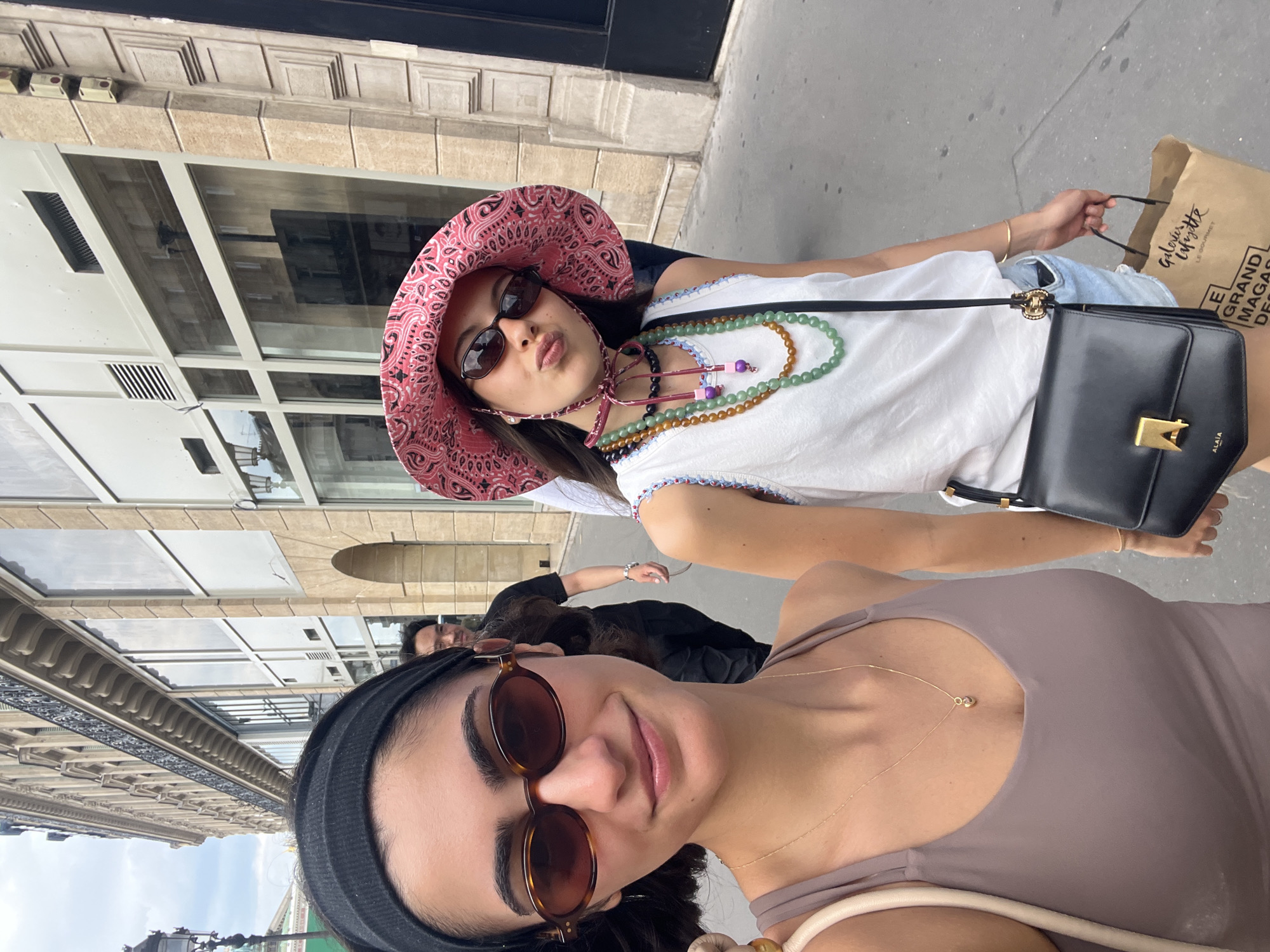 Two young women walk down a sidewalk in Paris, France, in front of a Haussmann-style building. The woman in the foreground on the left wears a black headband, sunglasses, and a taupe tank top with a delicate gold necklace. The woman on the right wears a pink bandana-print bucket hat, sunglasses, and a white sleeveless top with colorful beaded necklaces. She carries a black crossbody bag and is holding a shopping bag from Galeries Lafayette. In the background, a man walks away from the camera. The street is paved with cobblestones.