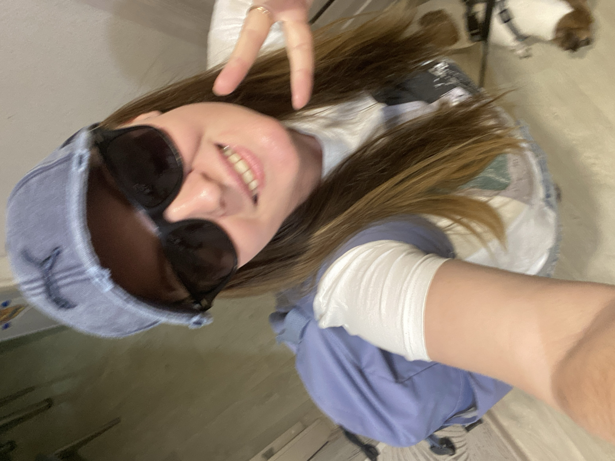 A person with long brown hair, wearing a denim baseball cap and sunglasses, smiles at the camera and makes a peace sign. They appear to be wearing a white long-sleeved shirt under a blue garment, possibly a denim jacket or overalls. The background is a combination of interior surfaces with a hint of furniture. There is no discernible information to indicate the location is in the city of Noviy, Russia.