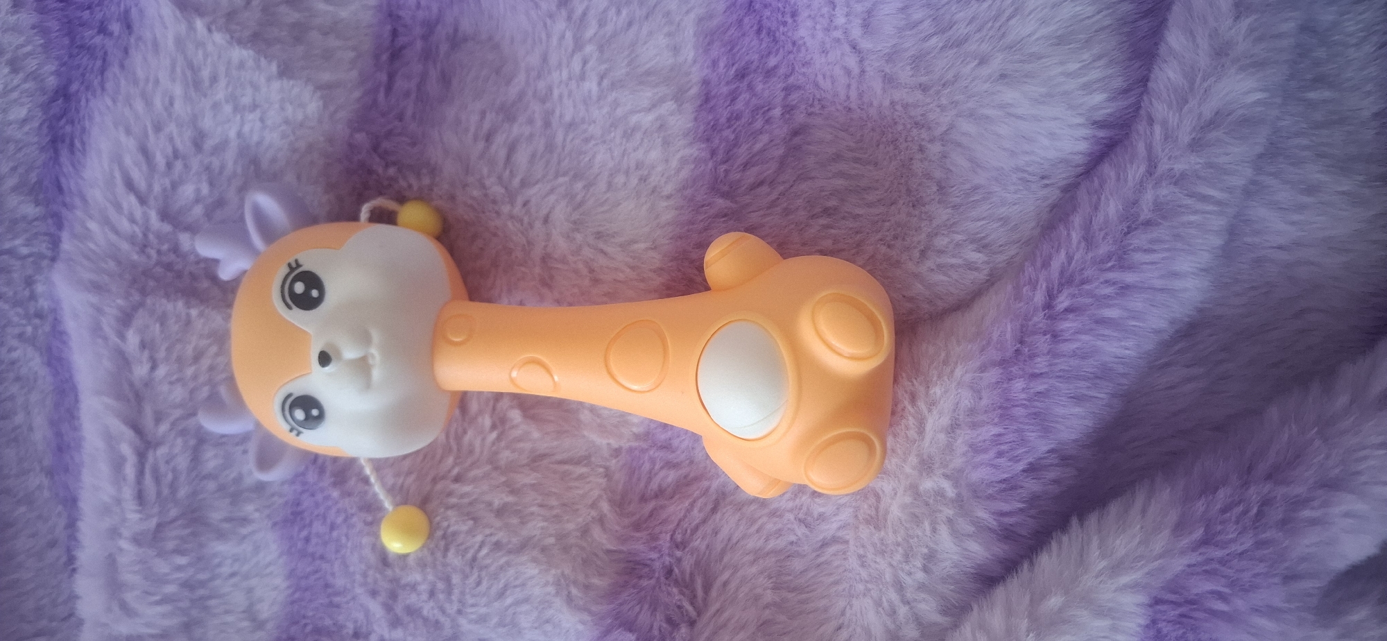 An orange giraffe-shaped rattle toy with purple antlers and yellow beads on white strings is lying on a purple and white striped furry blanket. The toy has a white oval on its body and circular patterns on its neck and legs. The background is a soft, plush furry blanket with stripes of light and dark purple. The scene is a close-up shot, with the toy positioned in the center of the frame. There is no indication of a city, location, or specific environment within Ust'-Katav, Russia.