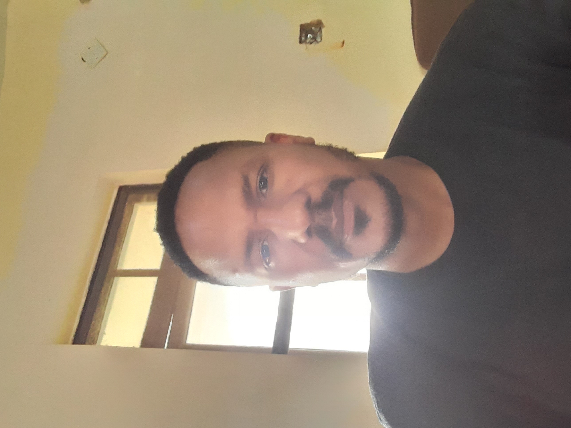 A medium shot of a Black man with dark hair and a beard, facing the camera, is set against a light-colored interior wall. He is wearing a dark-colored T-shirt. A window with a light-colored frame is visible behind him, letting in bright sunlight. The image was taken in an indoor setting in Gura Top, Nigeria.