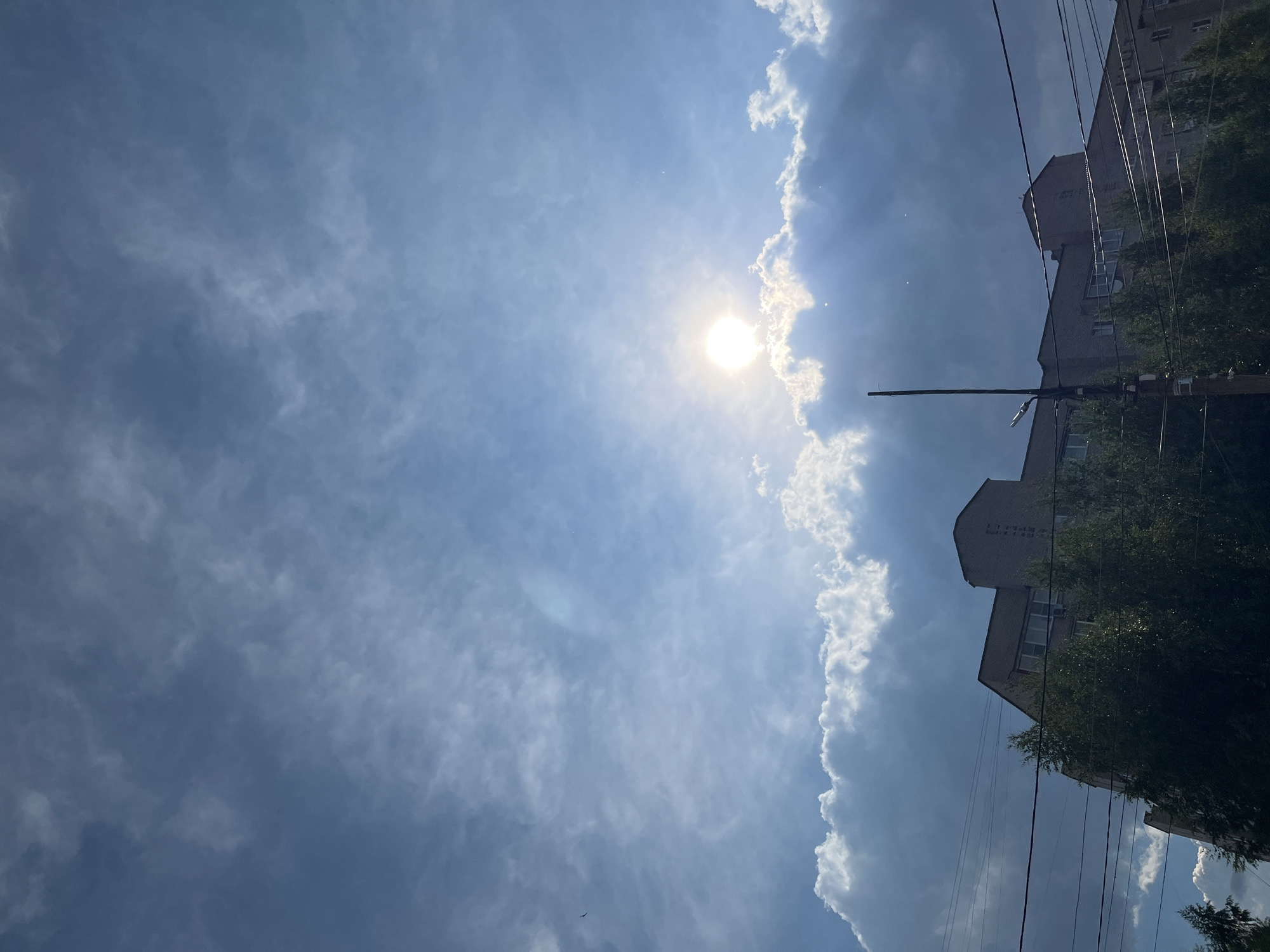 The image shows a view of the sky and buildings in Chaikovsky, Russia. The sky is blue with white and gray clouds, and the sun is shining brightly through the clouds. Below the sky, there are several buildings, some of which appear to be apartment buildings, and trees. There are also power lines visible in the foreground. The scene appears to be an outdoor urban environment.
