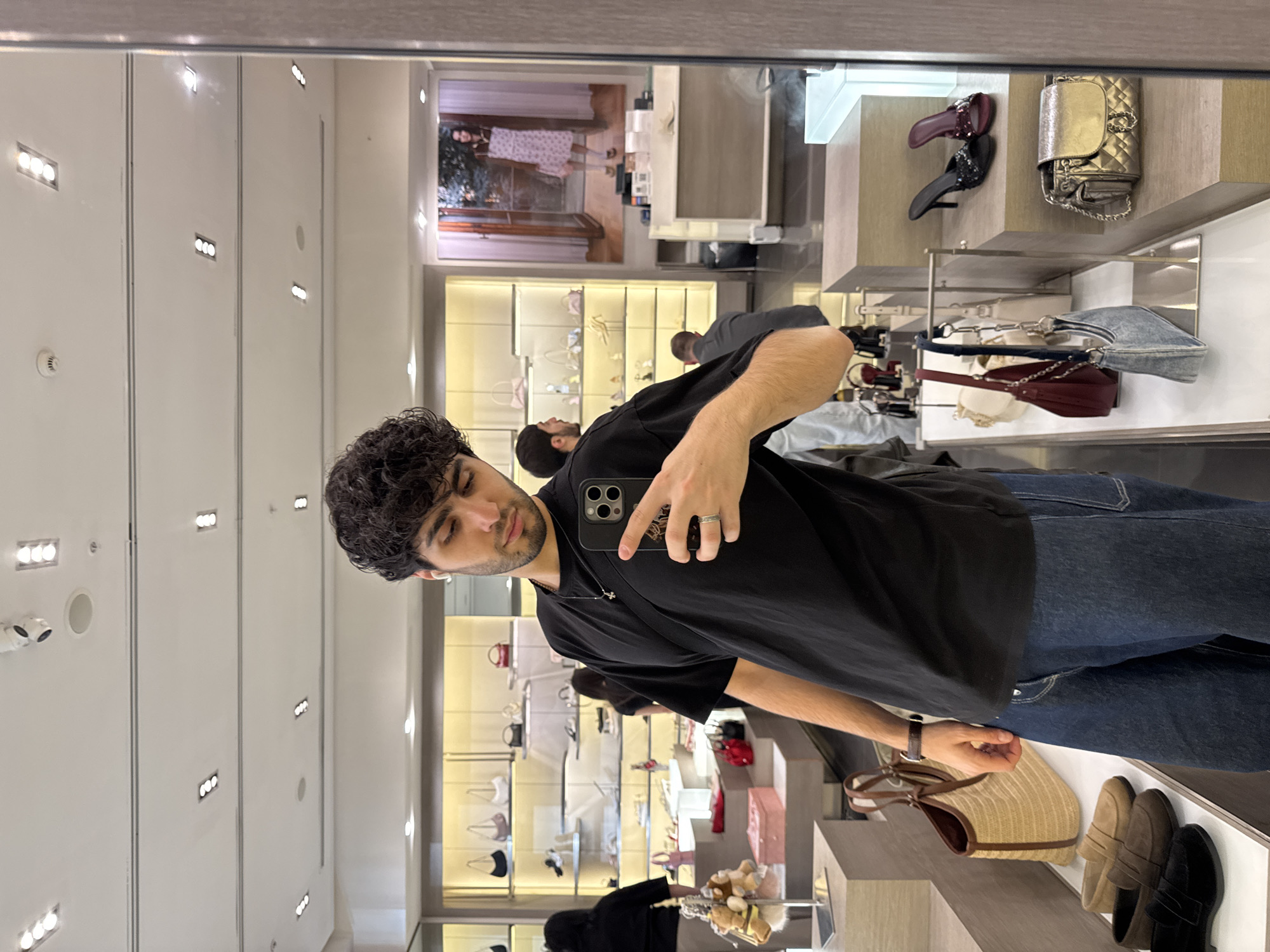 A man with curly dark hair and a beard takes a mirror selfie in what appears to be a retail store in Yerevan, Armenia. He is wearing a black t-shirt, dark jeans, and a watch on his left wrist, holding a smartphone with a prominent camera module in his right hand. His gaze is directed towards his phone, captured in the reflection.
The background reveals a well-lit interior with white walls and bright overhead lighting. Various display shelves are visible, showcasing an assortment of fashion accessories. Notable items include several handbags, such as a gold quilted bag, a denim bag, a red crescent-shaped bag, and a straw tote. A variety of shoes are also displayed, including burgundy and black heels, and brown and black loafers. Reflections of other customers or staff can be seen in the background, along with a large framed image or poster of a woman in a dress, adding to the store's ambiance. Security cameras are mounted on the ceiling. The overall scene suggests a modern fashion boutique or a department store section. No discernible text is clearly visible on any products, signs, or the man's phone case.