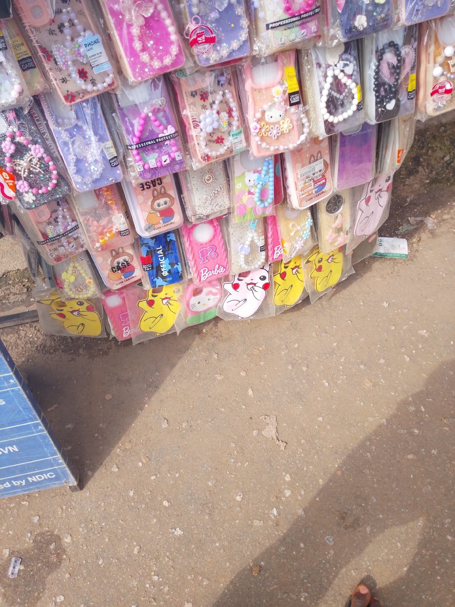 A street vendor displays an assortment of colorful phone cases for sale. The cases are arranged in rows and appear to be made from various materials like silicone, plastic, and fabric, with embellishments such as beads, glitter, and character designs like Hello Kitty and Pikachu. Some cases are attached to beaded lanyards. The background is a rough, dusty surface, likely a sidewalk or street, with a shadow cast from an unseen object or person. A blue sign with white text is partially visible in the lower left corner. The lighting suggests it is daytime, possibly outdoors in a market or commercial area.
