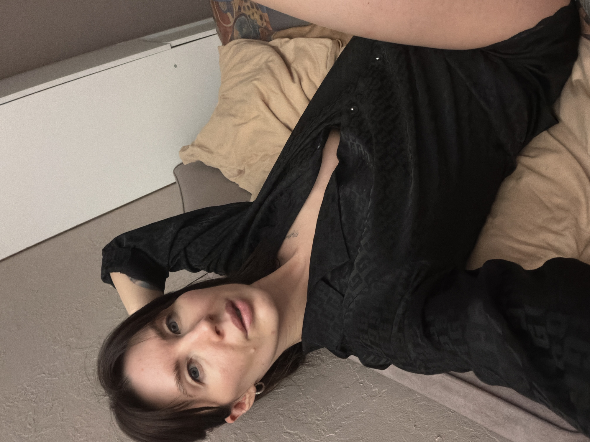A fair-skinned woman with dark hair lies on a bed with light tan pillows and a taupe-colored headboard. She is wearing a patterned black shirt, unbuttoned to reveal part of her chest. She is facing the camera. The location is Perm, Russia.