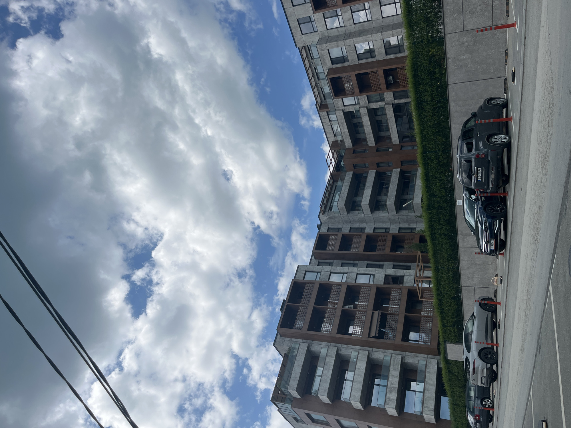 The image depicts an urban scene in Varna, Bulgaria. The buildings are modern, multi-story residential structures with a facade featuring a mix of gray and brown materials. The sky is partially cloudy with patches of blue visible. Several cars are parked along the street next to the building. Tall, green hedges line the street. Power lines are visible in the lower left corner of the image. Red and orange bollards mark parking spaces. The image has been rotated clockwise.