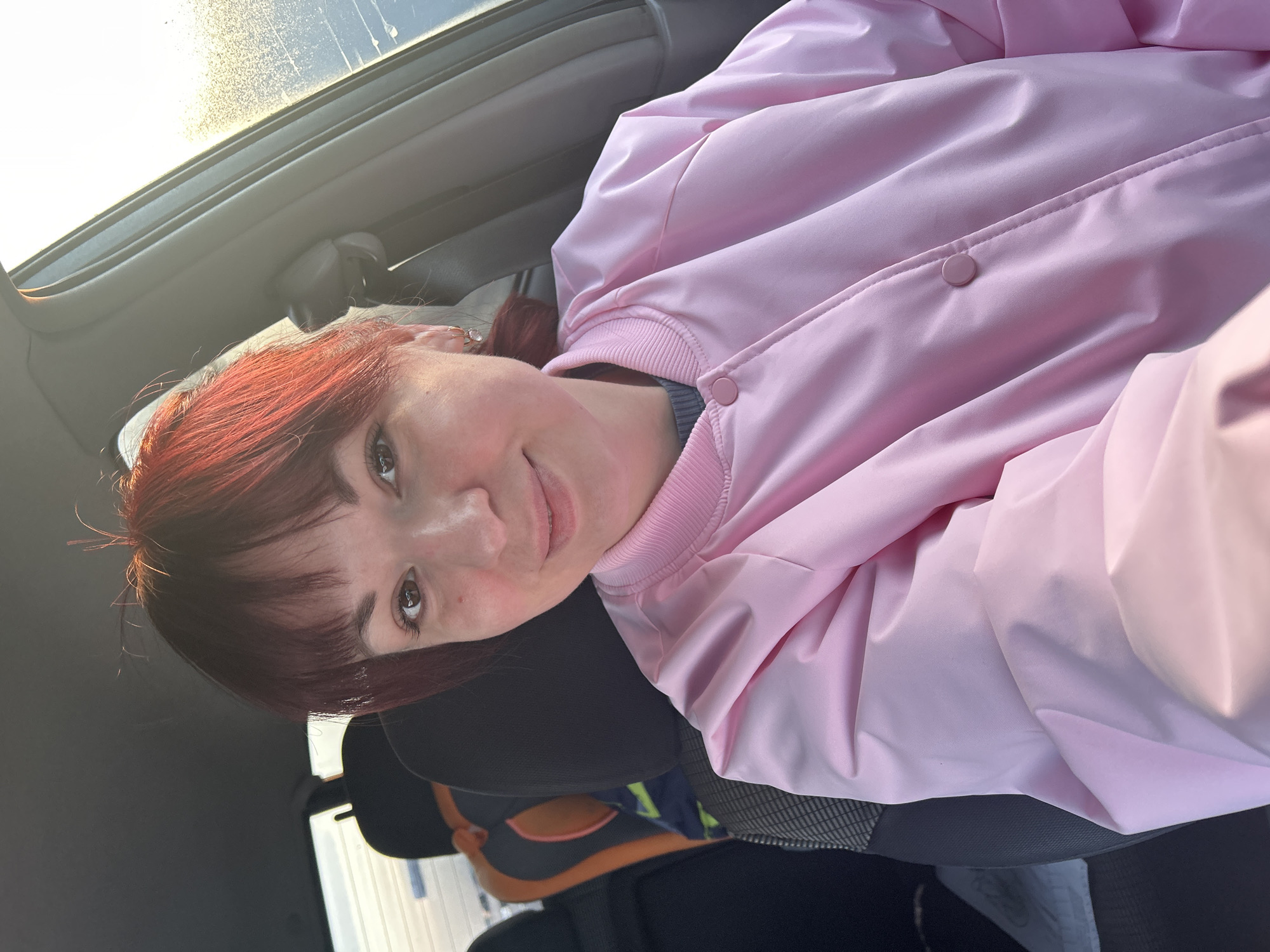 A person with red hair and a pink jacket is sitting in a car. The interior of the car is visible. The location is unspecified and cannot be determined from the image.
