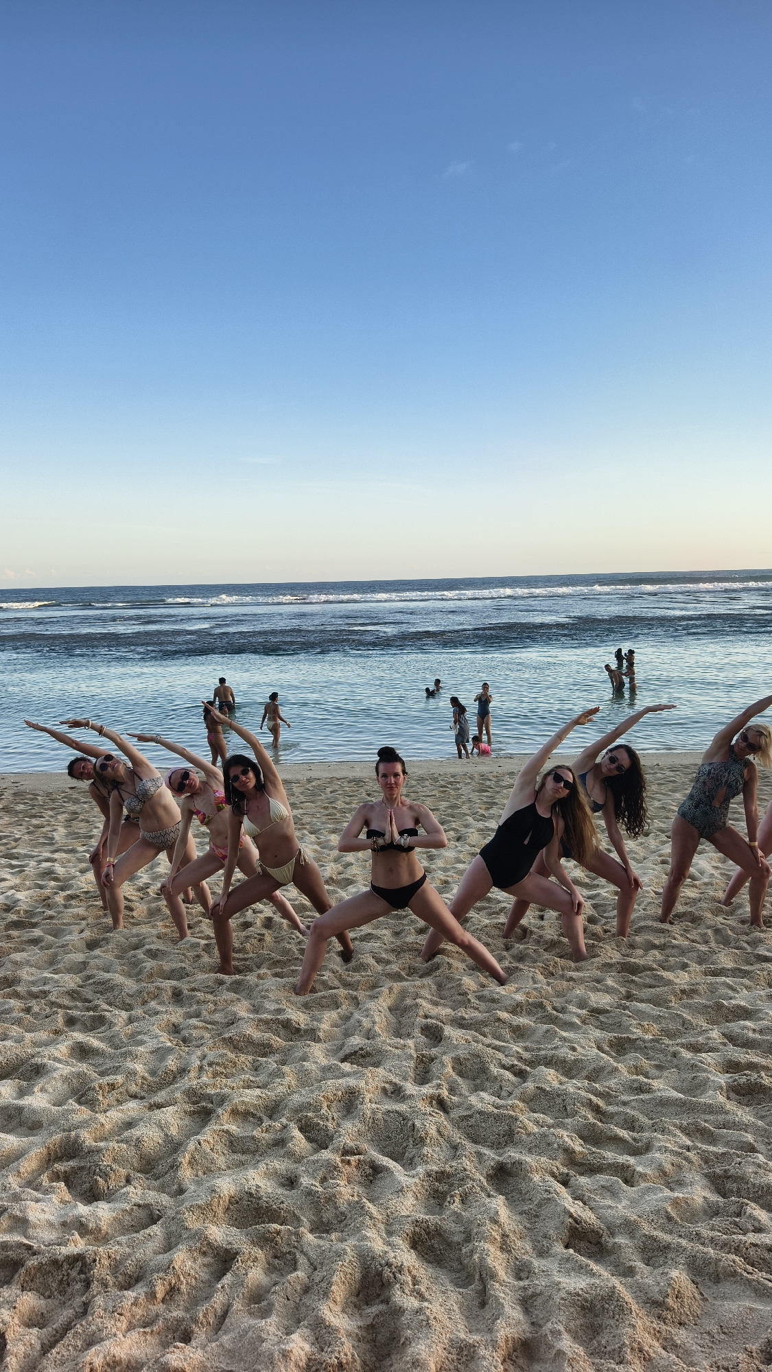 A group of women perform yoga on a beach in Kuta Selatan, Indonesia. They are on a sandy beach in bikinis. In the background is the ocean and several other people in the water. The sky is a clear blue.