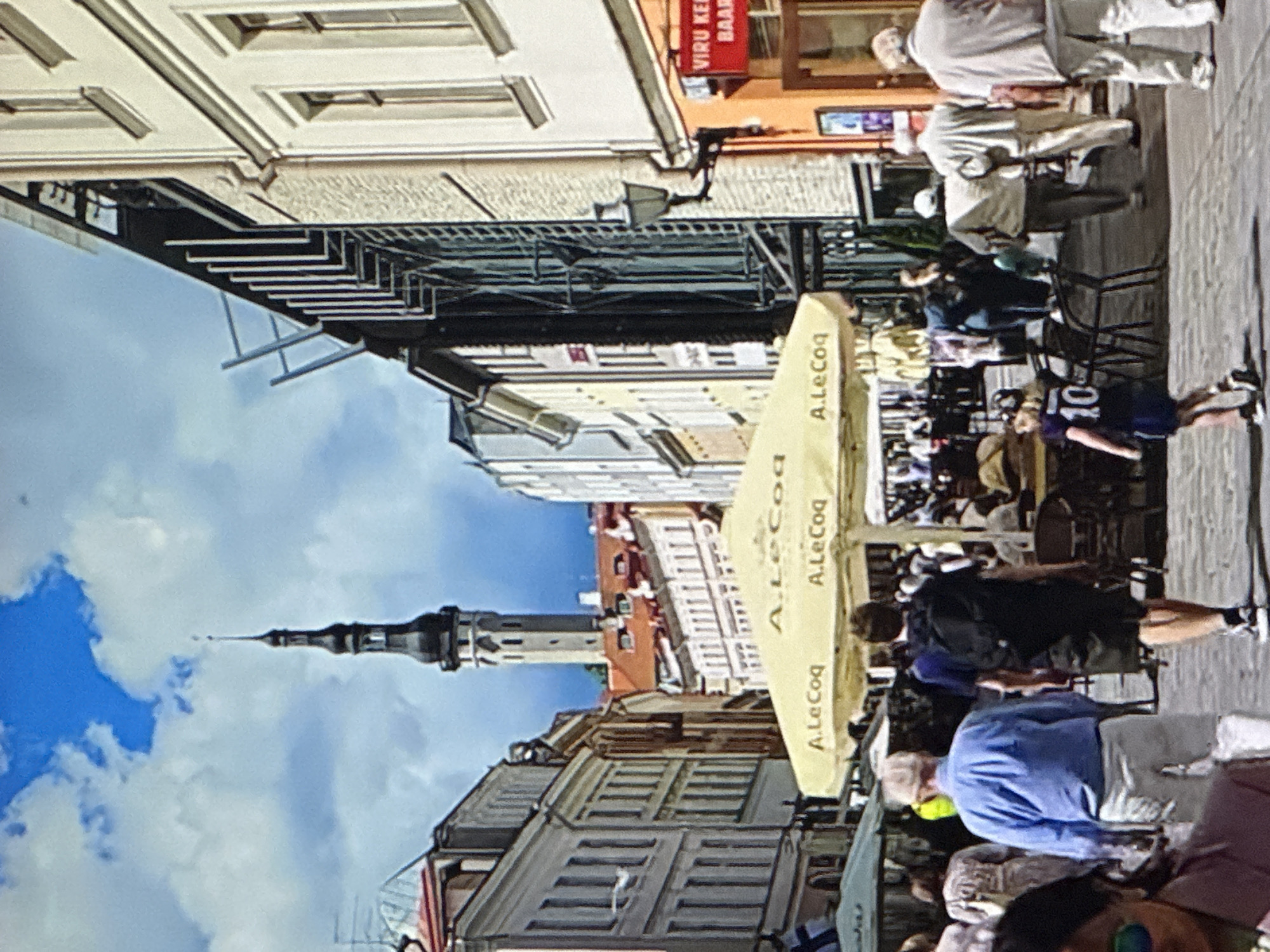 The image depicts a street scene in Tallinn, Estonia, likely in the Old Town, given the architecture and the prominent tower in the background, which resembles the St. Olaf's Church spire. It is a sunny day with scattered clouds in a blue sky. A narrow street is lined with multi-story buildings on either side, exhibiting traditional European architecture with various window styles and rooflines. In the foreground, a yellow umbrella with the "A. Le Coq" logo is prominently displayed, indicating an outdoor seating area for a cafe or restaurant. Several people are walking along the street. In the mid-ground, a man in a blue shirt and white shorts is walking away from the camera, followed by a person in a black backpack. A young child wearing a dark blue t-shirt with the number "10" on the back is walking ahead of the other pedestrians. In the background, there are more people seated at tables under umbrellas, and further down the street, a tall, slender tower with a dark spire dominates the skyline. The overall atmosphere suggests a lively, tourist-frequented area.