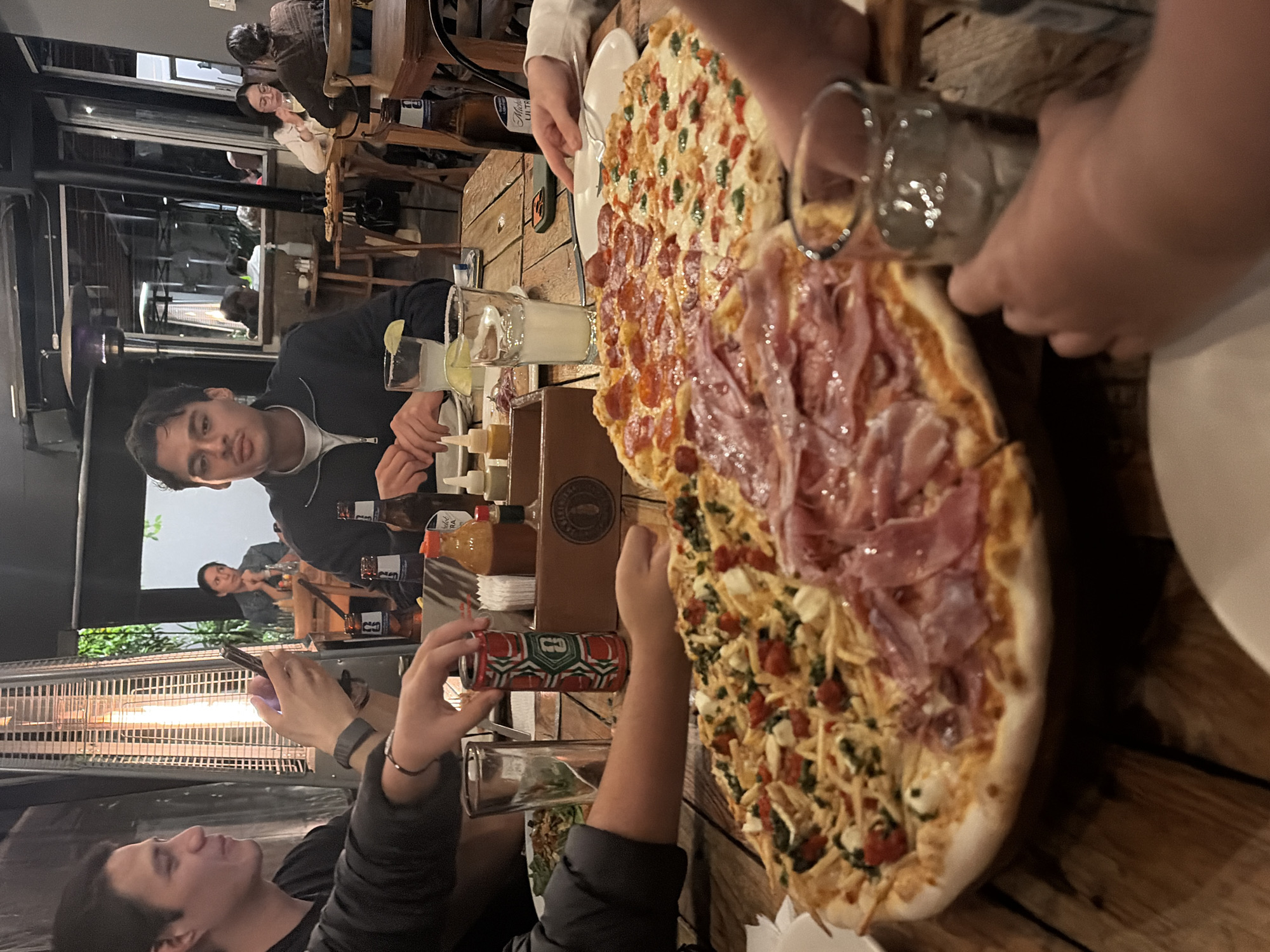 A tilted, indoor scene shows a group of people gathered around a large, multi-flavored pizza on a wooden table. The pizza has at least three distinct sections: one with pepperoni, one with ham, and another with a mix of vegetables and cheese. There are drinks, including glasses with ice and beverages in cans, also on the table.
At least three people are visible: one taking a picture with a phone, another seated and looking directly at the camera, and part of a third. In the background, there are glimpses of other people and parts of the indoor establishment, and what appears to be a patio heater. The location is in the city of Unknown, Unknown.