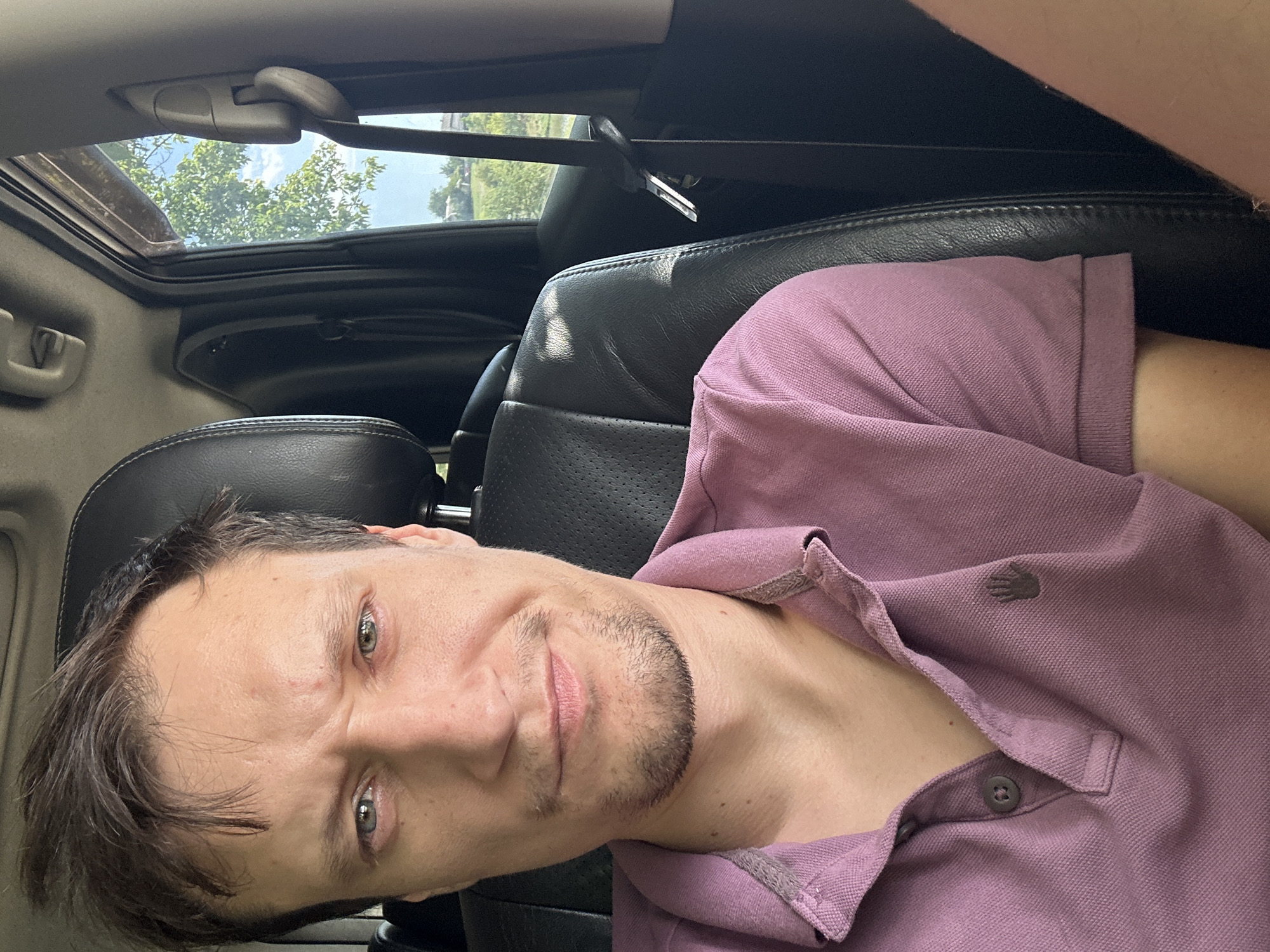 A close-up, eye-level selfie shows a man with brown hair and light green eyes looking directly at the camera. He has stubble on his chin and is wearing a light purple polo shirt with a small logo on the left side of his chest. The man is seated in the driver's seat of a vehicle, with the headrest of the passenger seat visible to his right. Sunlight streams in from the right side of the vehicle, illuminating the scene. Through the car window, a green tree and a blue sky with white clouds are visible, suggesting an outdoor setting. The scene appears to be a residential area outside of the city of Makiv, Ukraine, possibly a rural or suburban landscape.